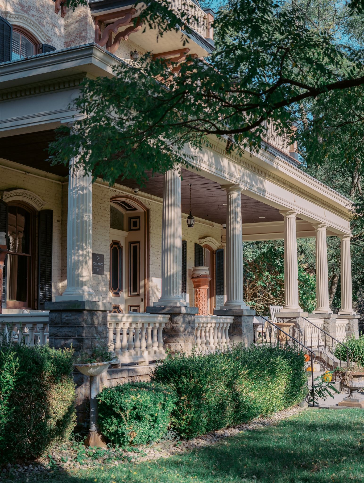 Front porch of a large, historic house with tall white columns, decorative railing, black shutters, and lush greenery. The house has a brick and painted exterior with a staircase leading up to the entrance.