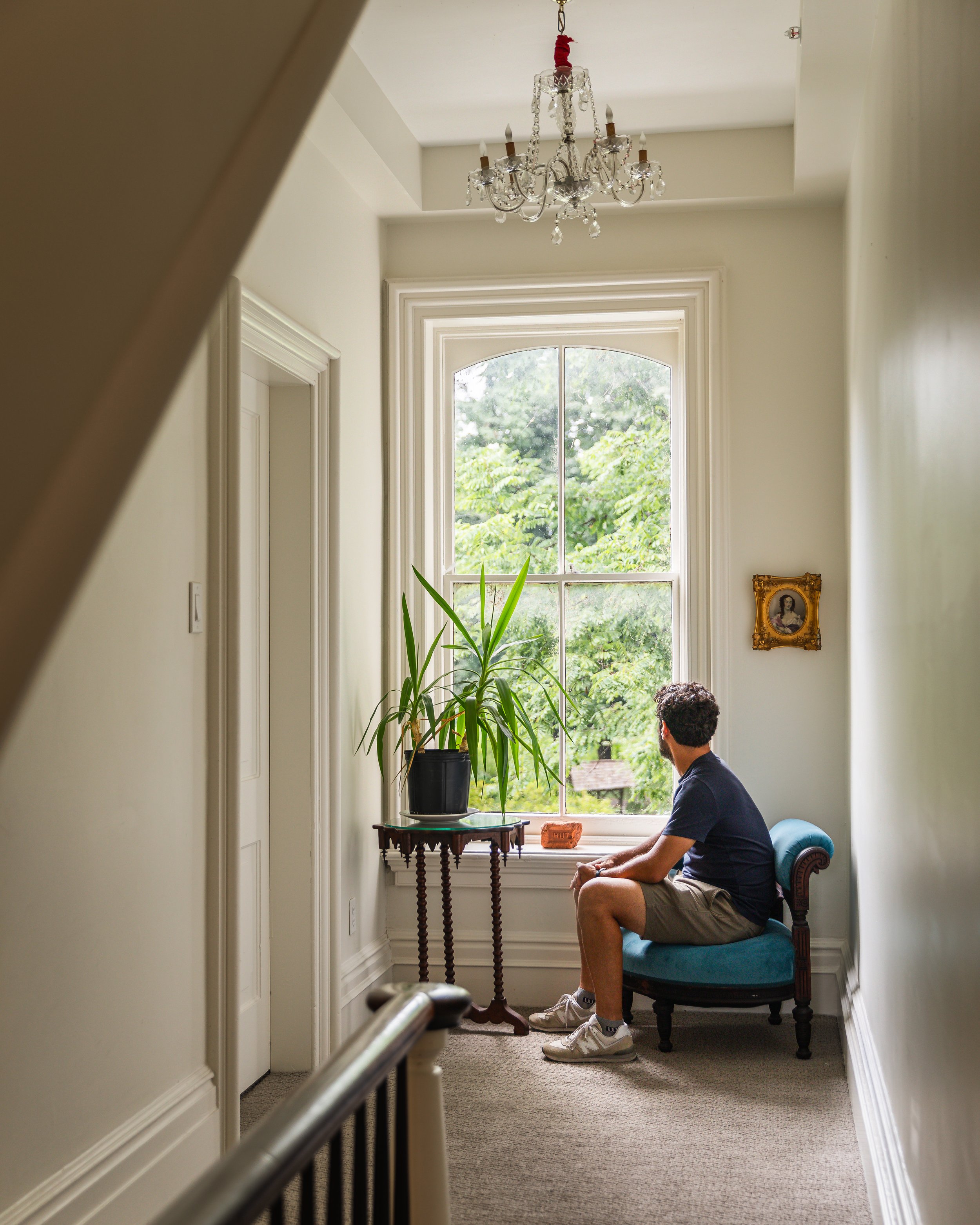 A man sitting on a vintage armchair near a large window with lush greenery outside in a bright, elegant hallway, with a small table holding a potted plant and a brick-shaped item, and a chandelier hanging from the ceiling.
