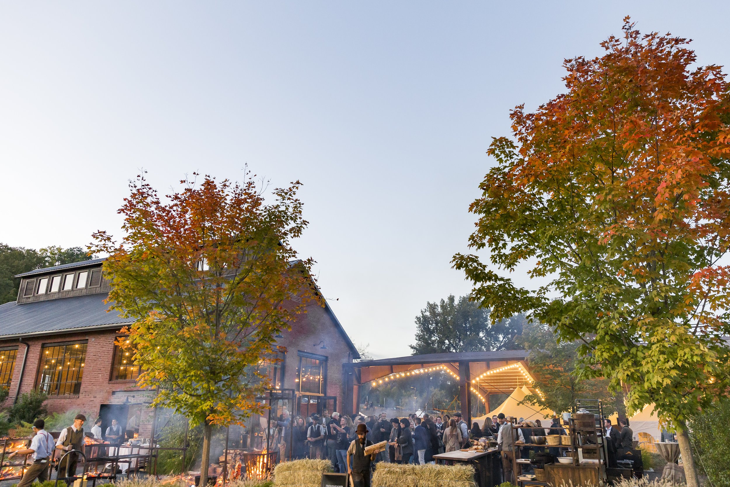 Outdoor evening gathering at a rustic barn with string lights, people socializing near a campfire, and fall foliage with orange and green leaves.