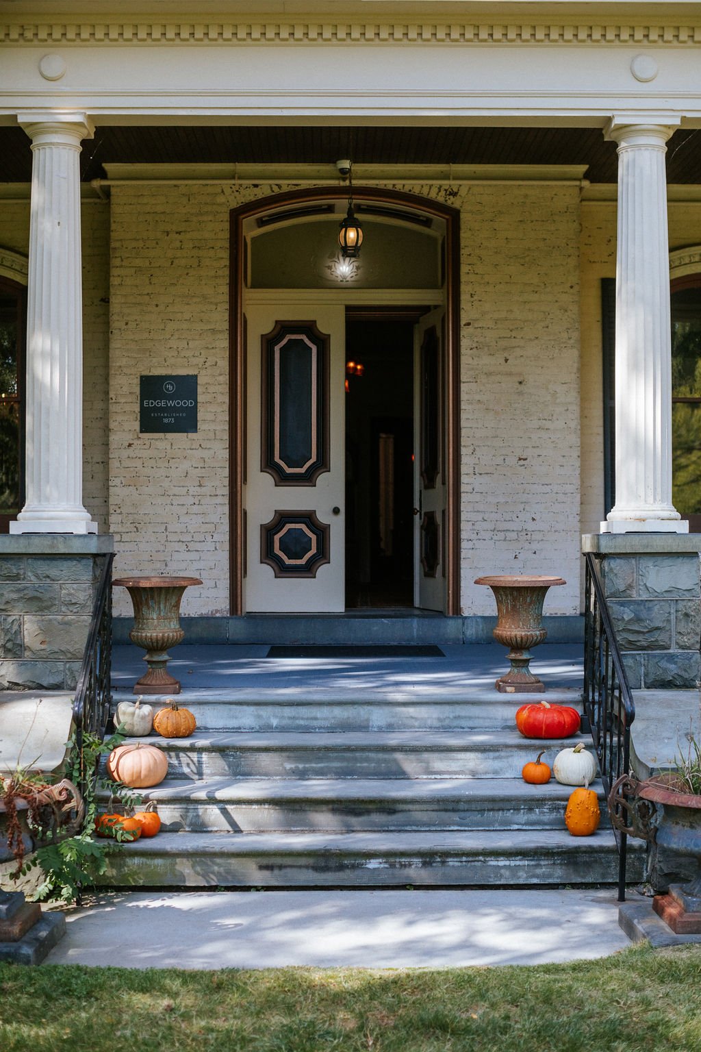 Front porch of a vintage house decorated with pumpkins for fall, featuring white and orange pumpkins placed on the steps and planters. The house has brick walls, tall white columns, and a door with intricate wood details.