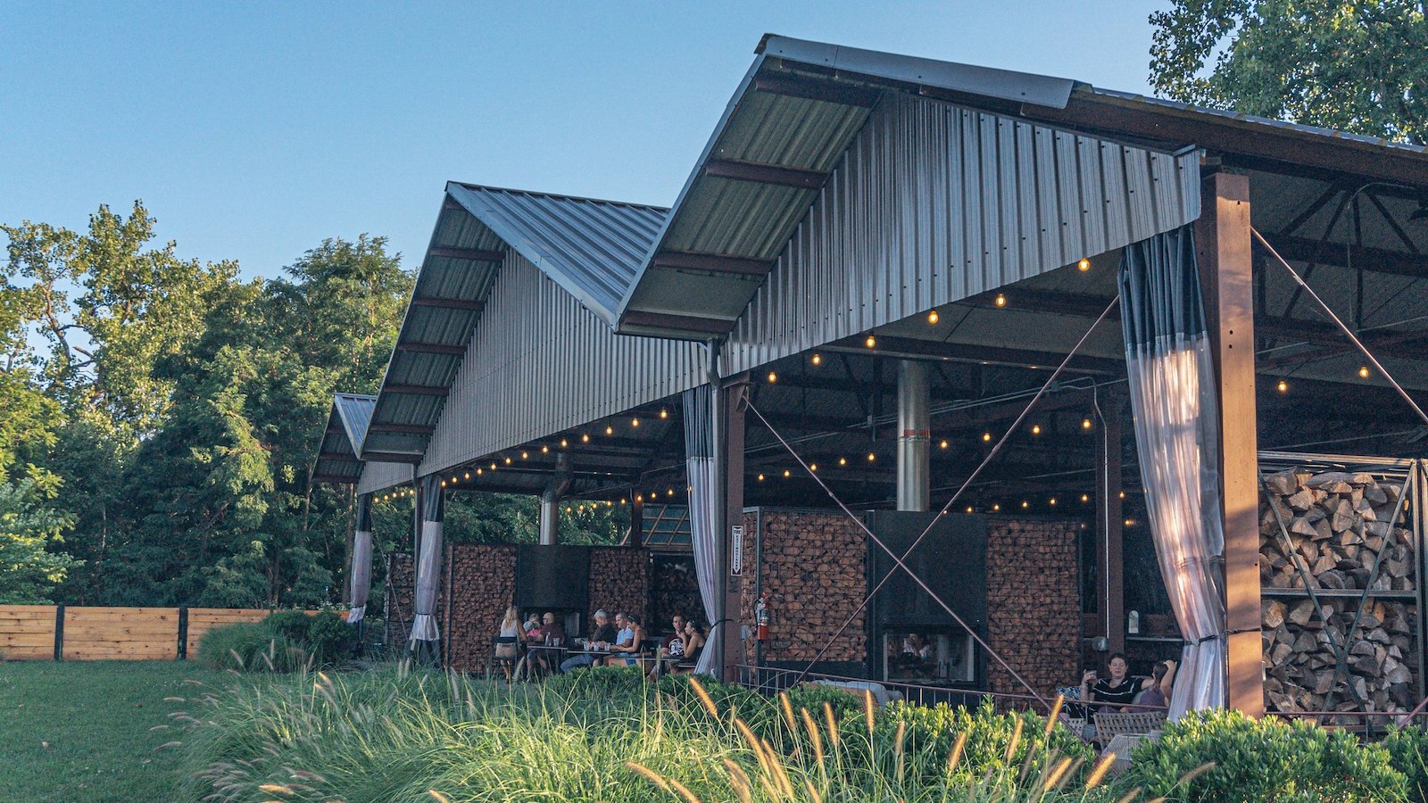 Outdoor pavilion with people sitting at tables, wood firewood stacked on the sides, string lights hanging, and trees in the background during daylight.
