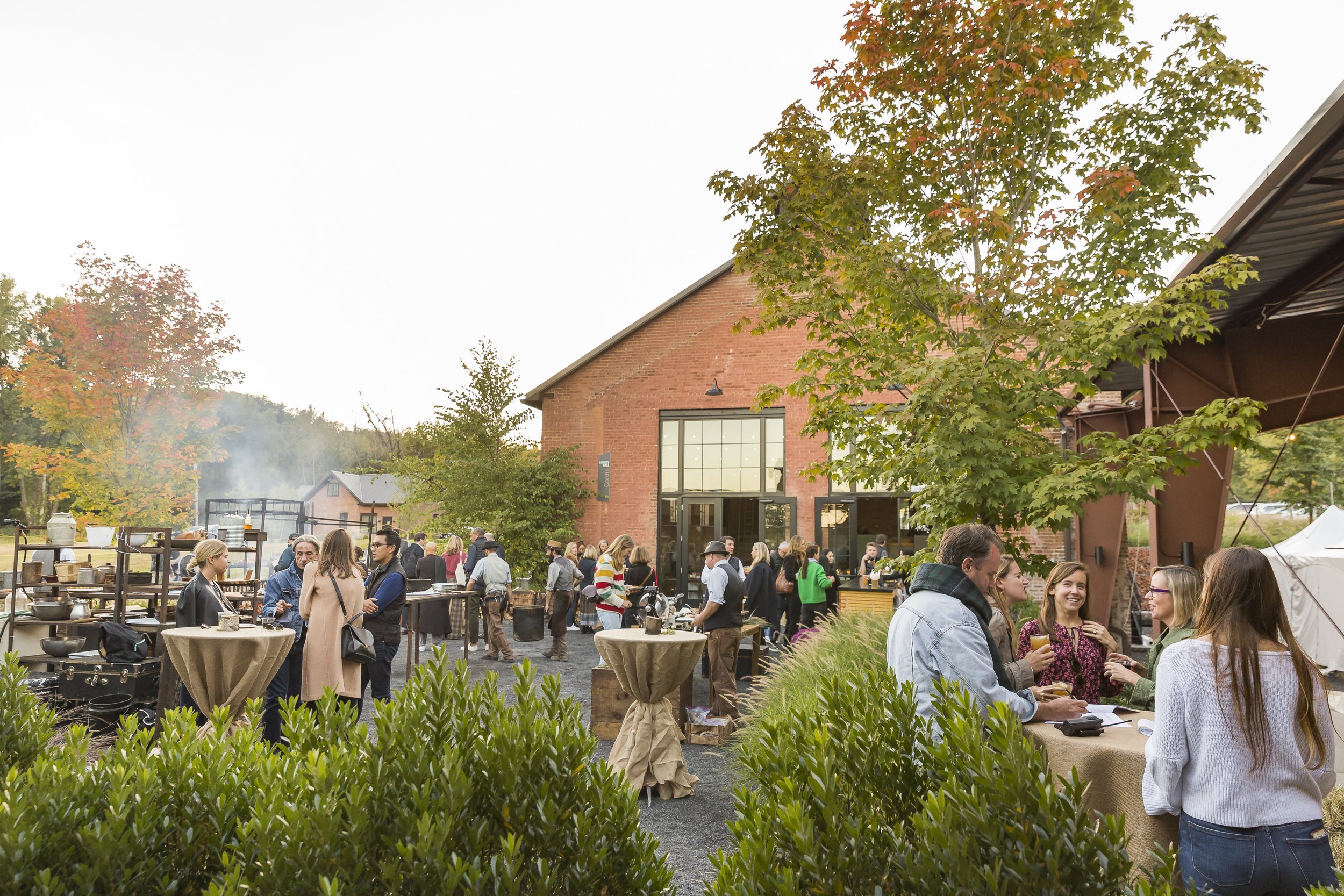 People attending an outdoor event at a rustic venue with brick and metal architecture, surrounded by trees and greenery, during fall.