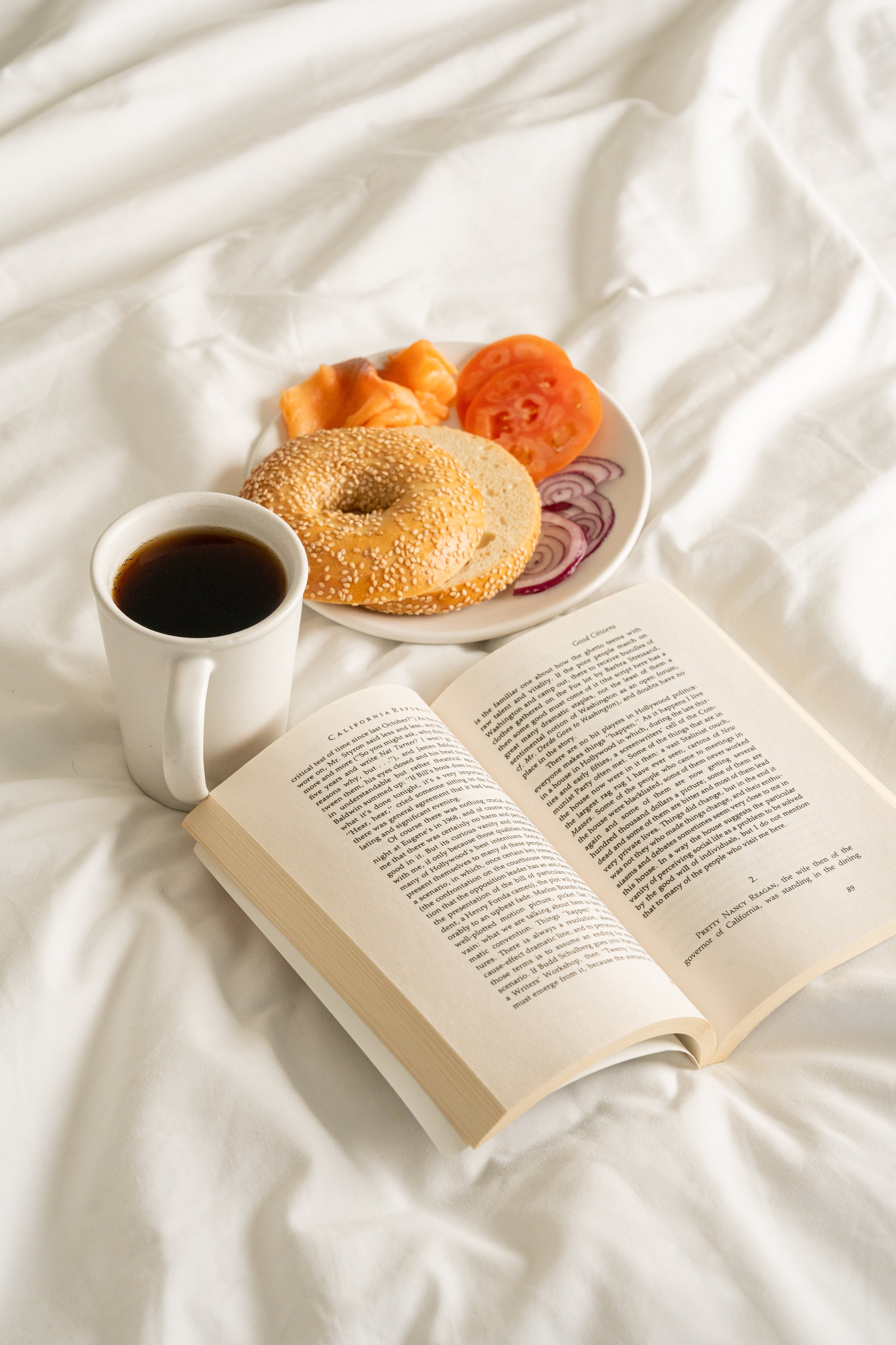 Open book next to a cup of coffee, a plate with a sesame bagel, tomato slices, onion slices, and vacuum-packed salmon on a white bed sheet.