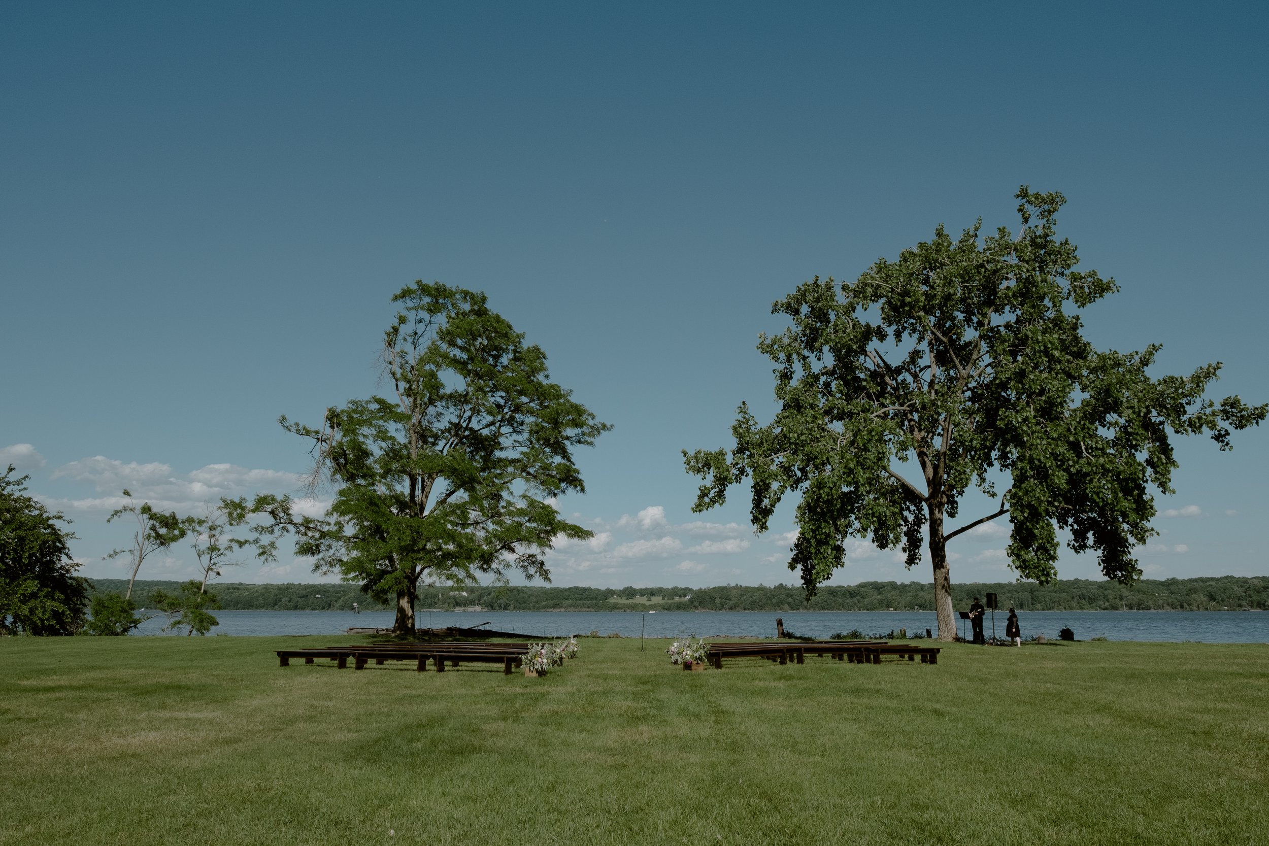 A grassy area with two large trees near a body of water, with a clear blue sky and a few clouds, and two people near the trees.