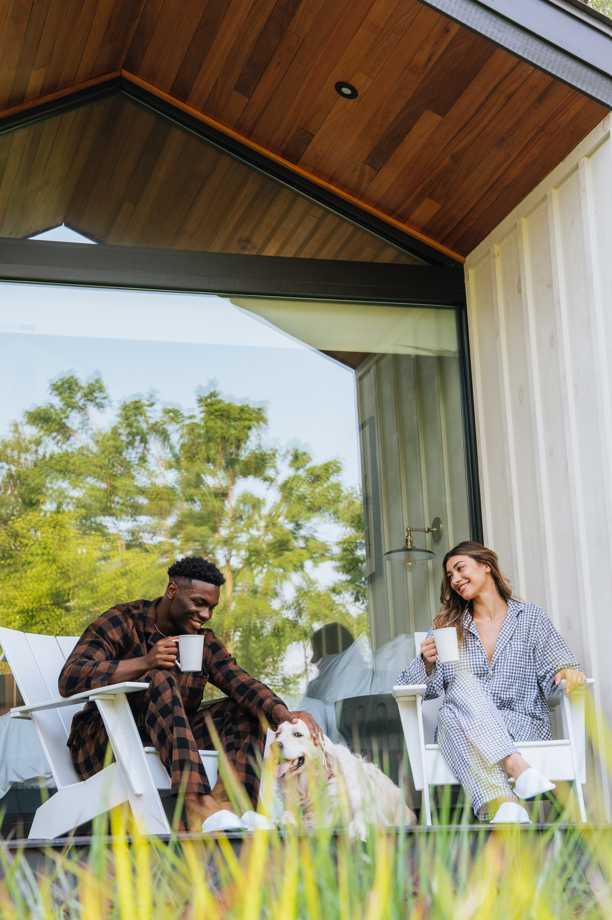 A man and woman sitting outside on chairs with a dog, enjoying drinks, on a porch with a wooden ceiling and large glass sliding door, with greenery in the background.
