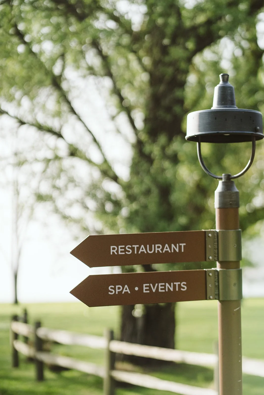 Signpost with arrows pointing to a restaurant, spa, and events, in an outdoor setting with trees and a wooden fence in the background.