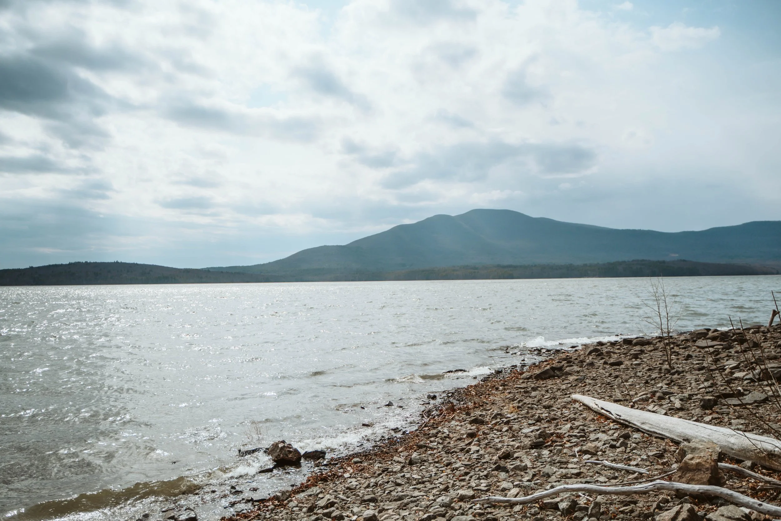 View of the Hudson River with a rocky shoreline, distant hills or mountains, and a cloudy sky.