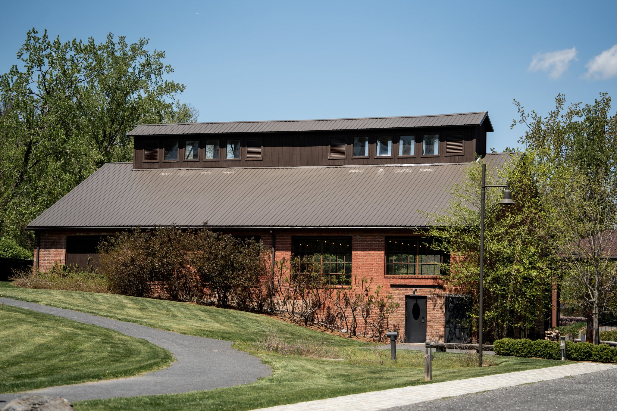 A brick house with a sloped metal roof and a small upper window structure, surrounded by green trees and a grassy lawn under a blue sky.