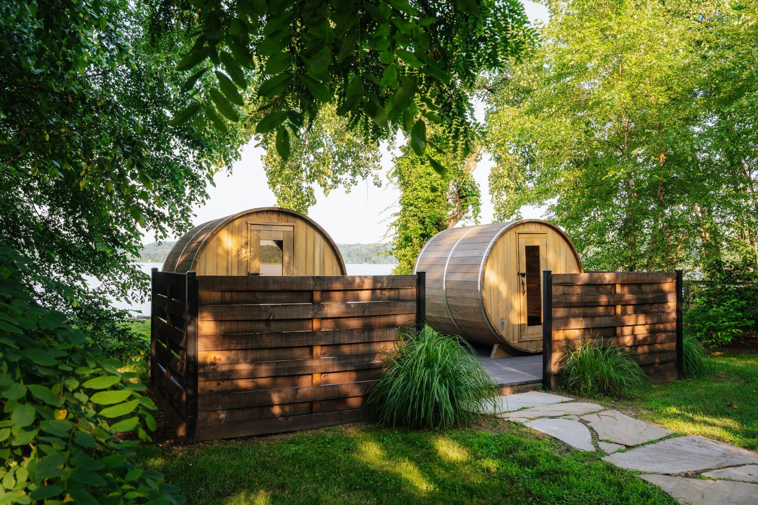Two wooden, waterfront saunas, surrounded by lush green trees and a fenced yard, near the Hudson River. 