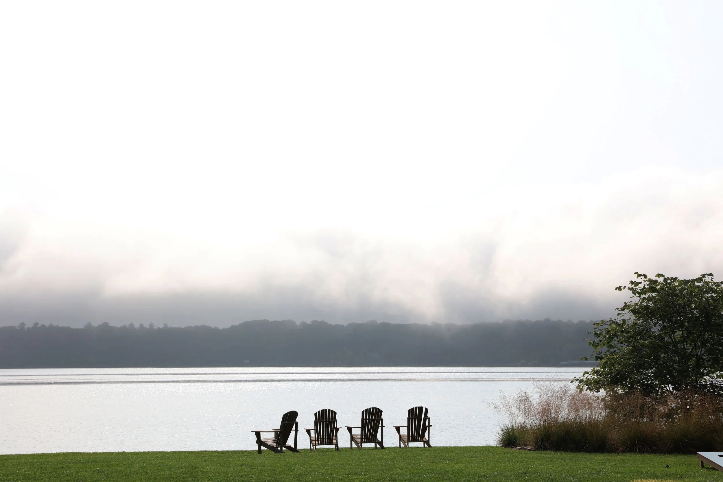 Six Adirondack chairs on a lawn overlooking the Hudson River with mountains in the distance under a cloudy sky.