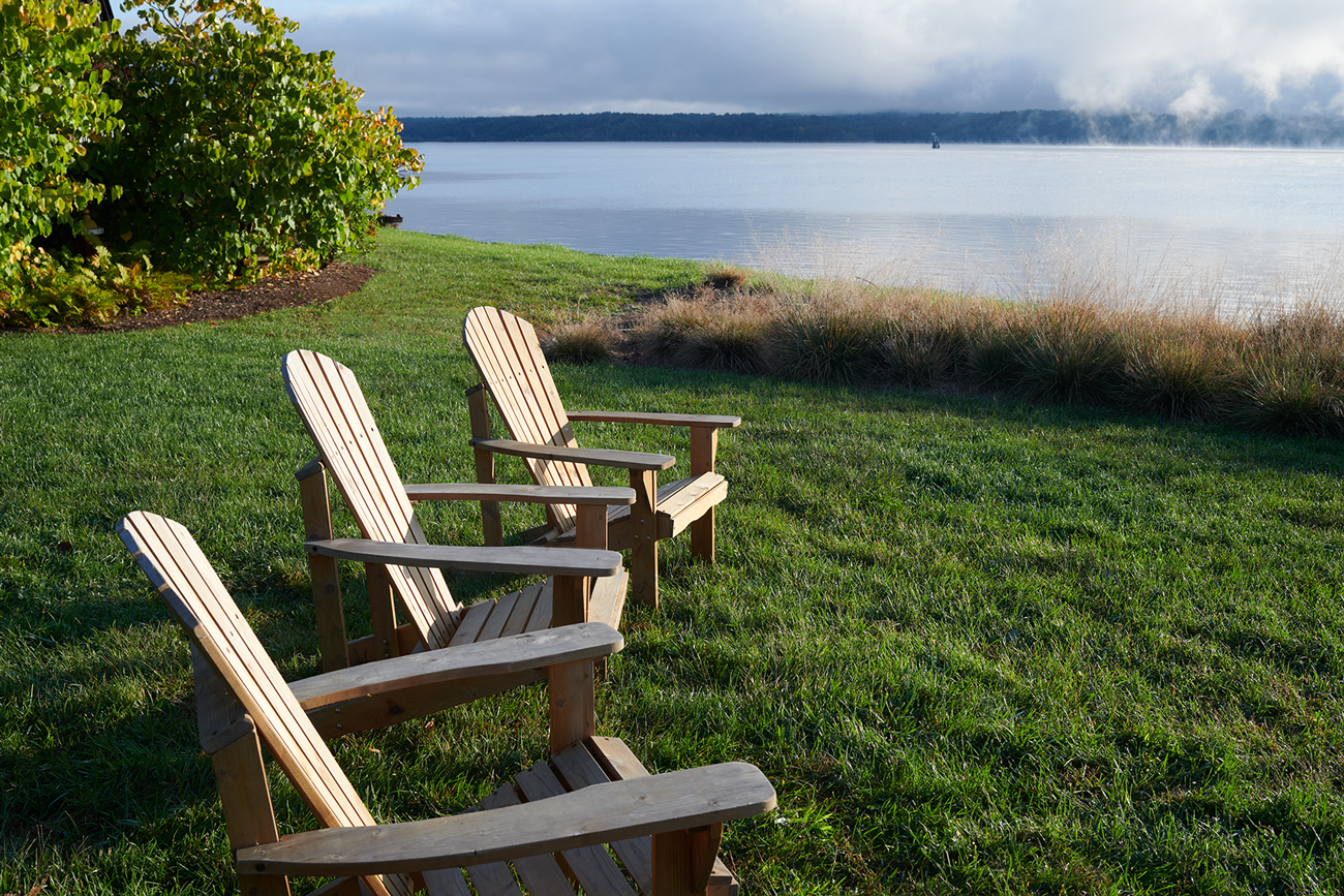 Three Adirondack chairs facing a calm body of water with grass, bushes, and distant hills under a partly cloudy sky.