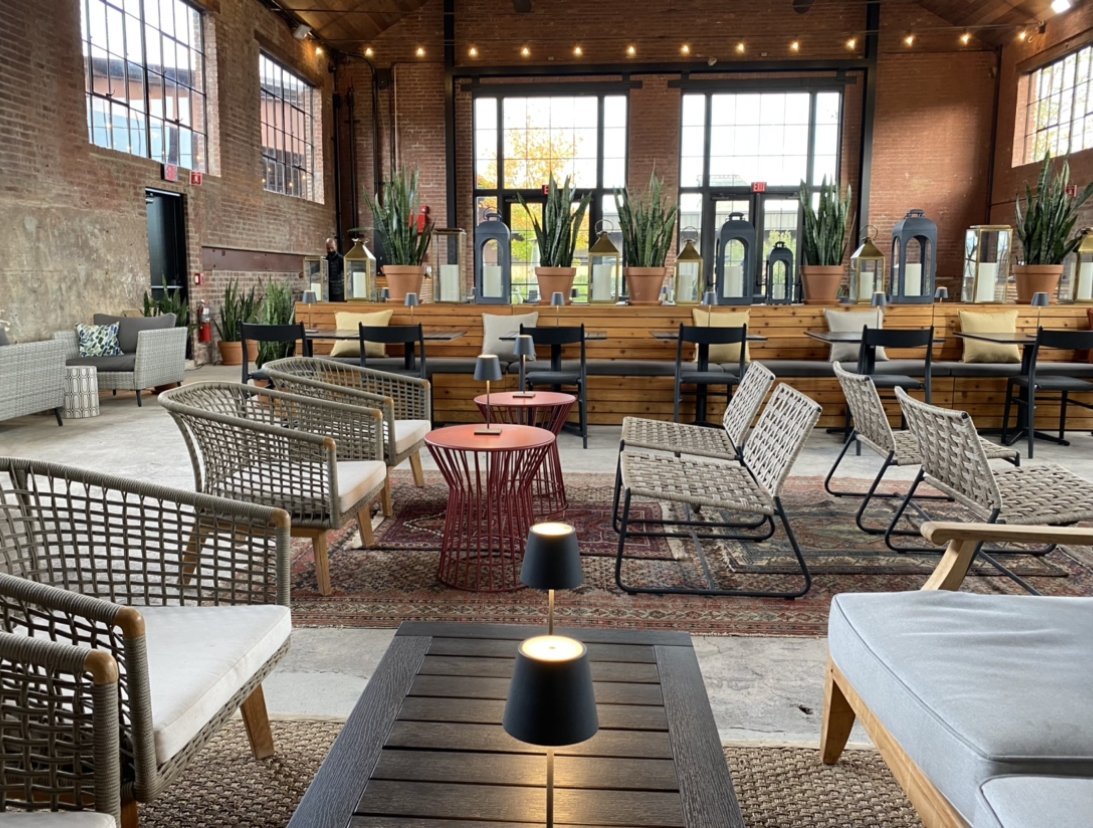 An indoor lounge area with multiple chairs, small tables, potted plants, and large windows letting in natural light.