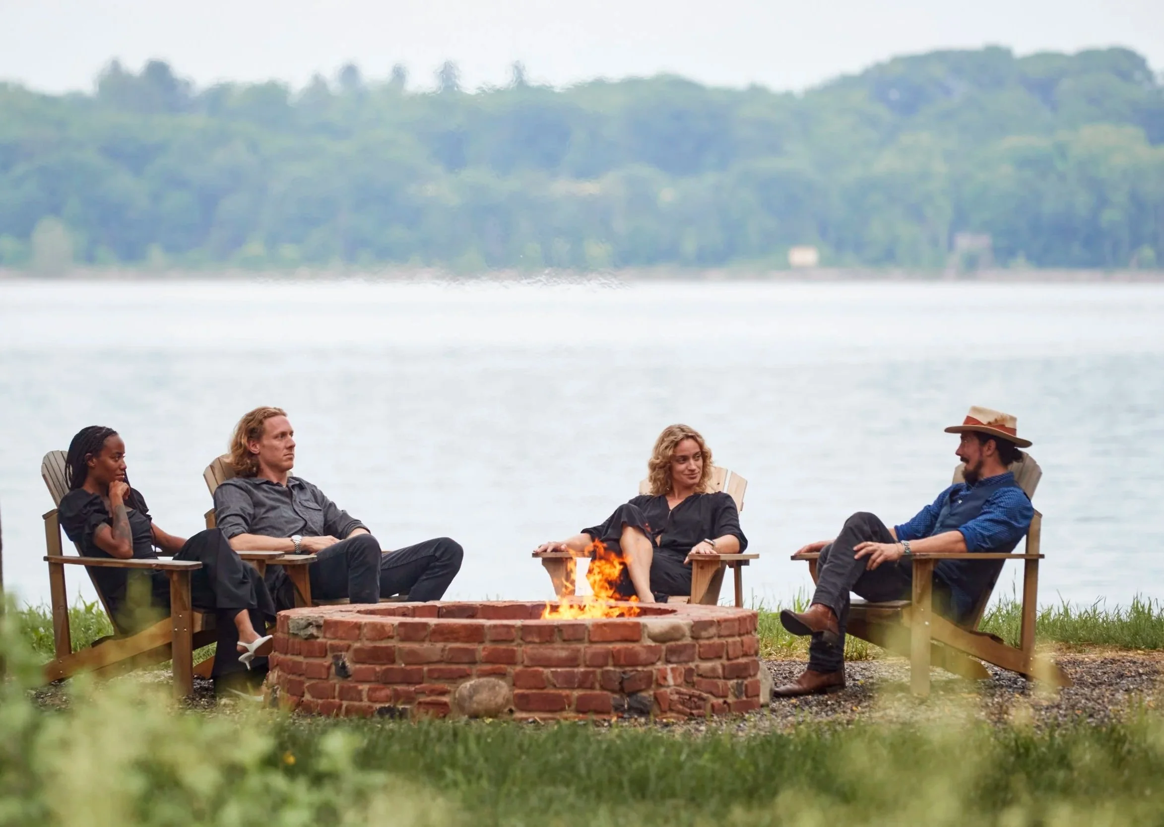 Four people sitting around a campfire near the Hudson riverfront, with a forested landscape in the background, engaging in conversation.