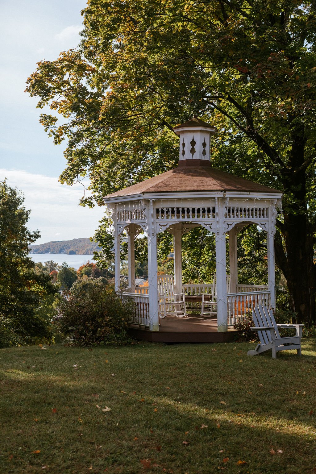 A white gazebo with a brown roof and decorative trim, surrounded by green trees and grass, with a view of a body of water and distant trees in the background.