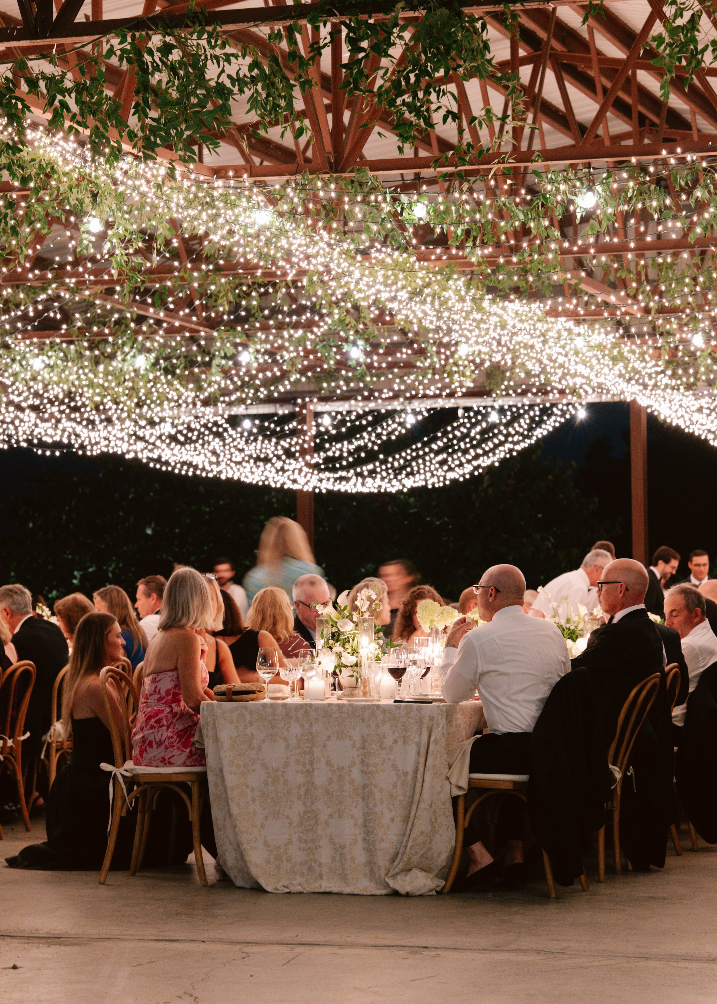 People dining at a celebration under string lights with greenery overhead at night