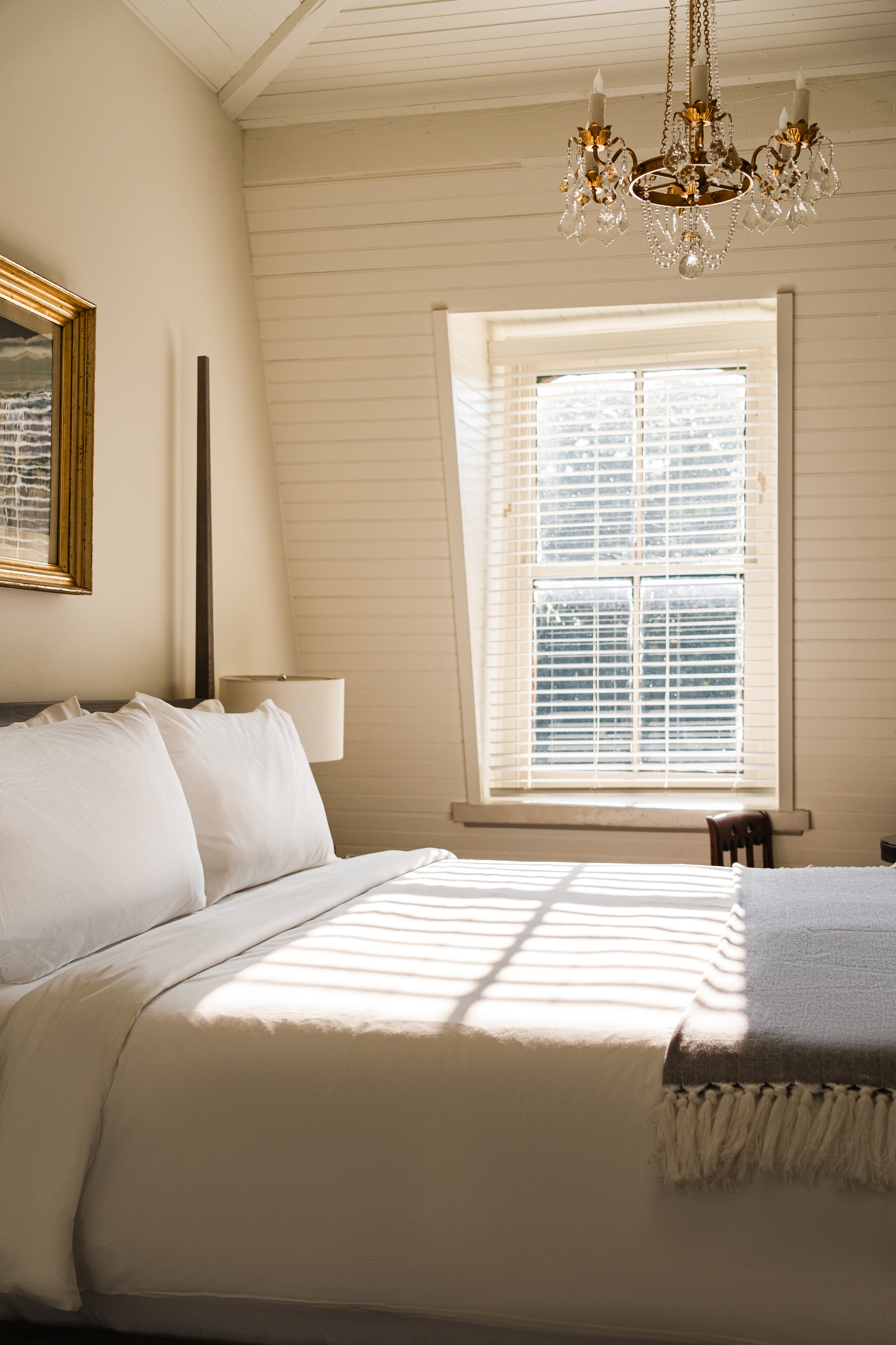 A bedroom with a bed, three pillows, and a blanket, illuminated by natural sunlight through a window with blinds, and a chandelier hanging from the ceiling.