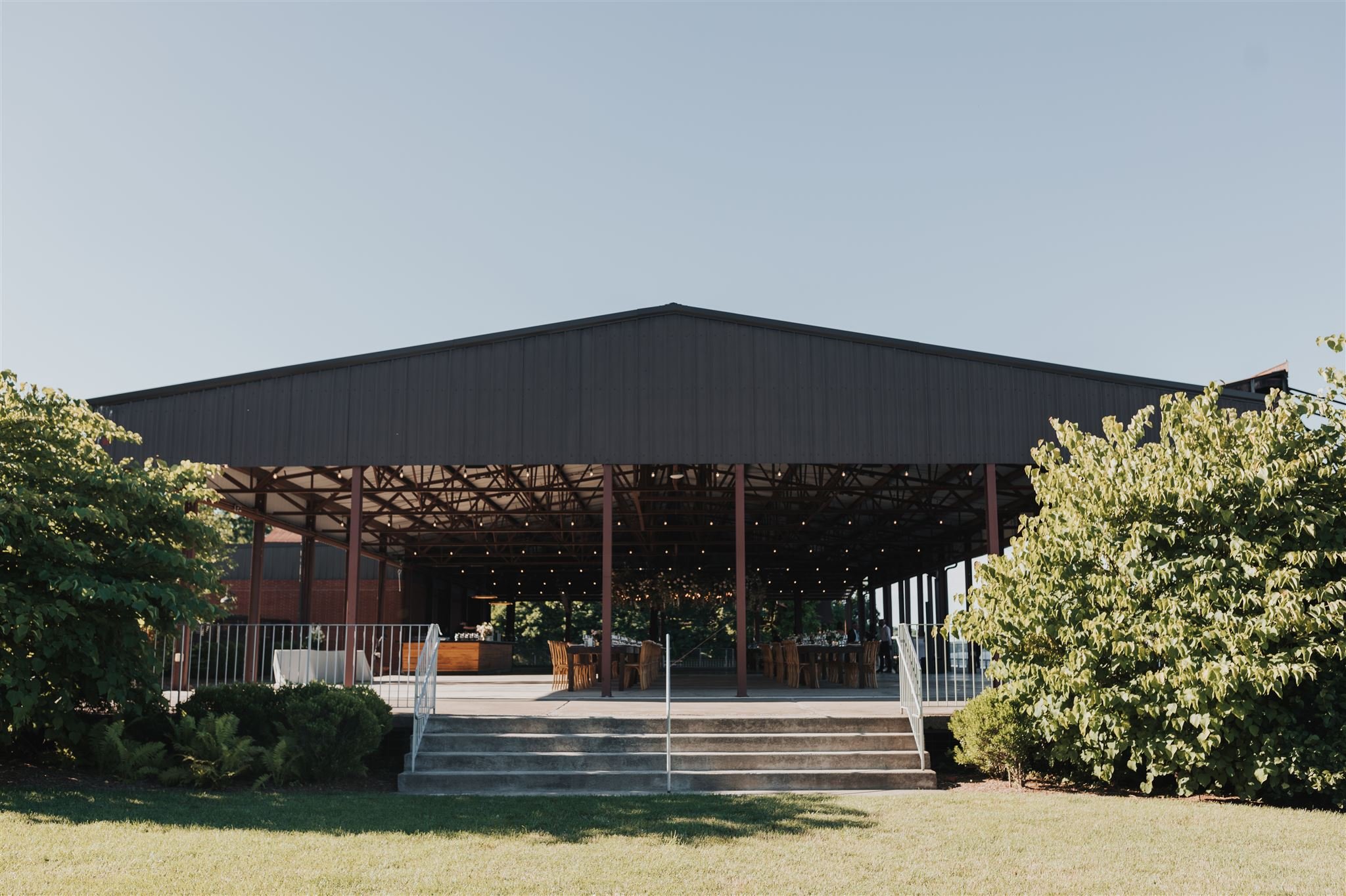 Outdoor event pavilion with black roof and string lights, set on a grassy area with trees. It has a stairway and tables inside.