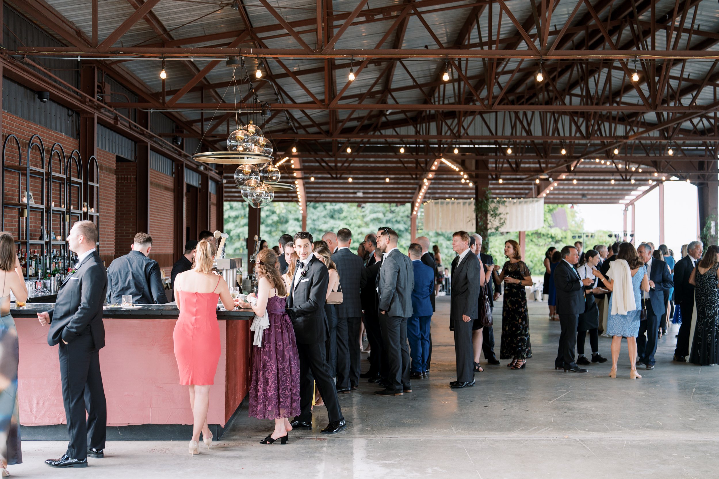 Guests at a social event or wedding reception in an outdoor pavilion, standing in line at a bar with string lights hanging from the ceiling, and greenery visible in the background.