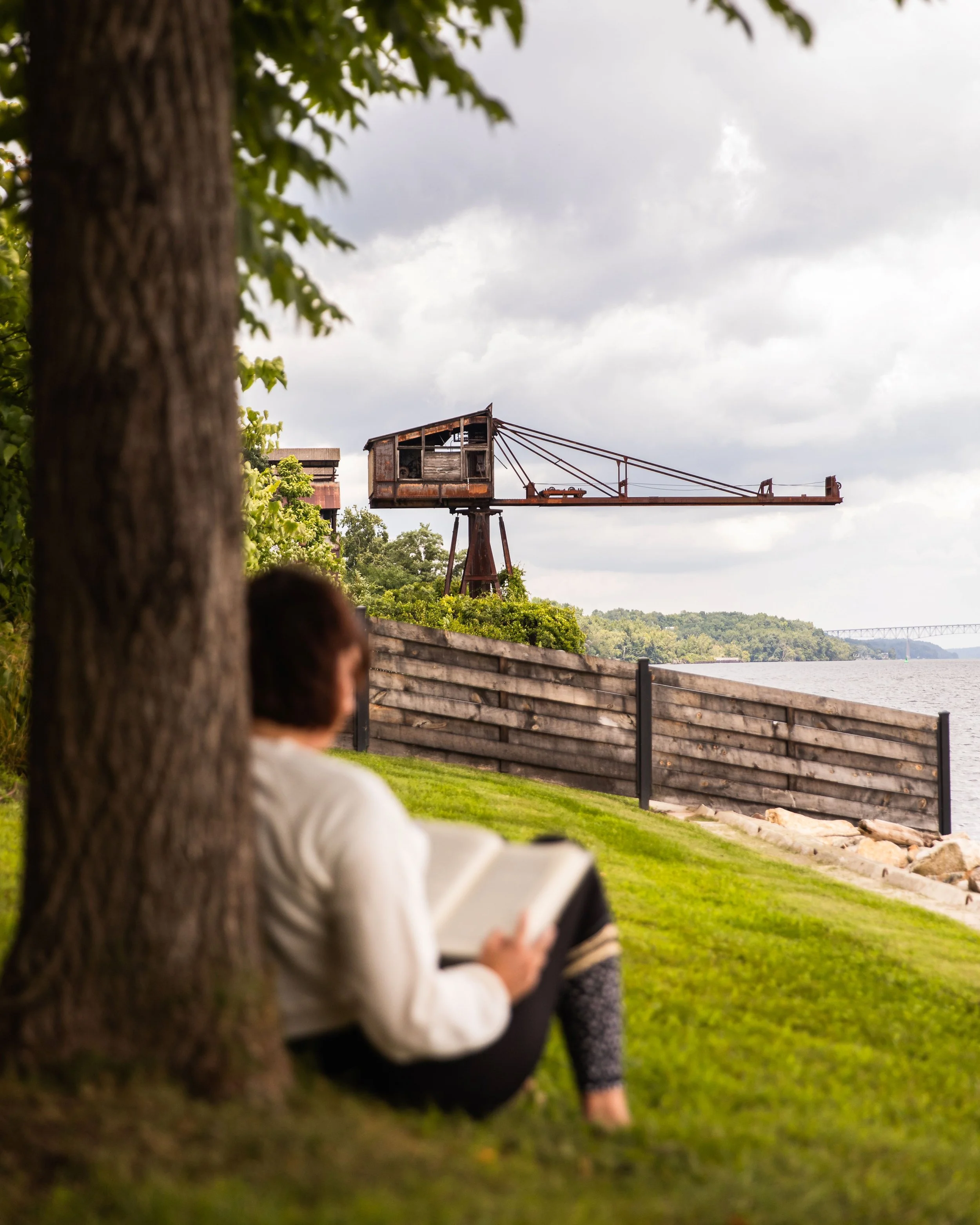 A person sitting under a tree reading a book near the Hudson River with the historic, Hutton Brickyard crane in the background.