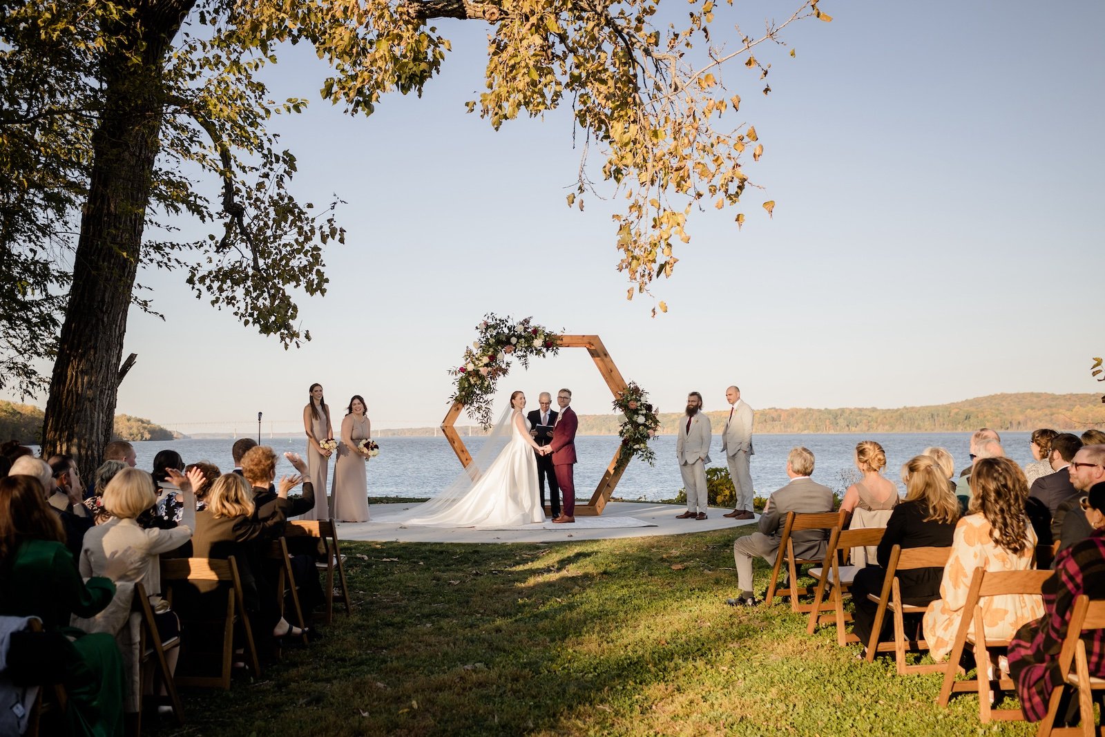 A couple getting married outdoors by a lake with guests seated on both sides. The bride and groom are holding hands and facing each other under a hexagonal wooden arch decorated with flowers, while an officiant conducts the ceremony. The scene is sun