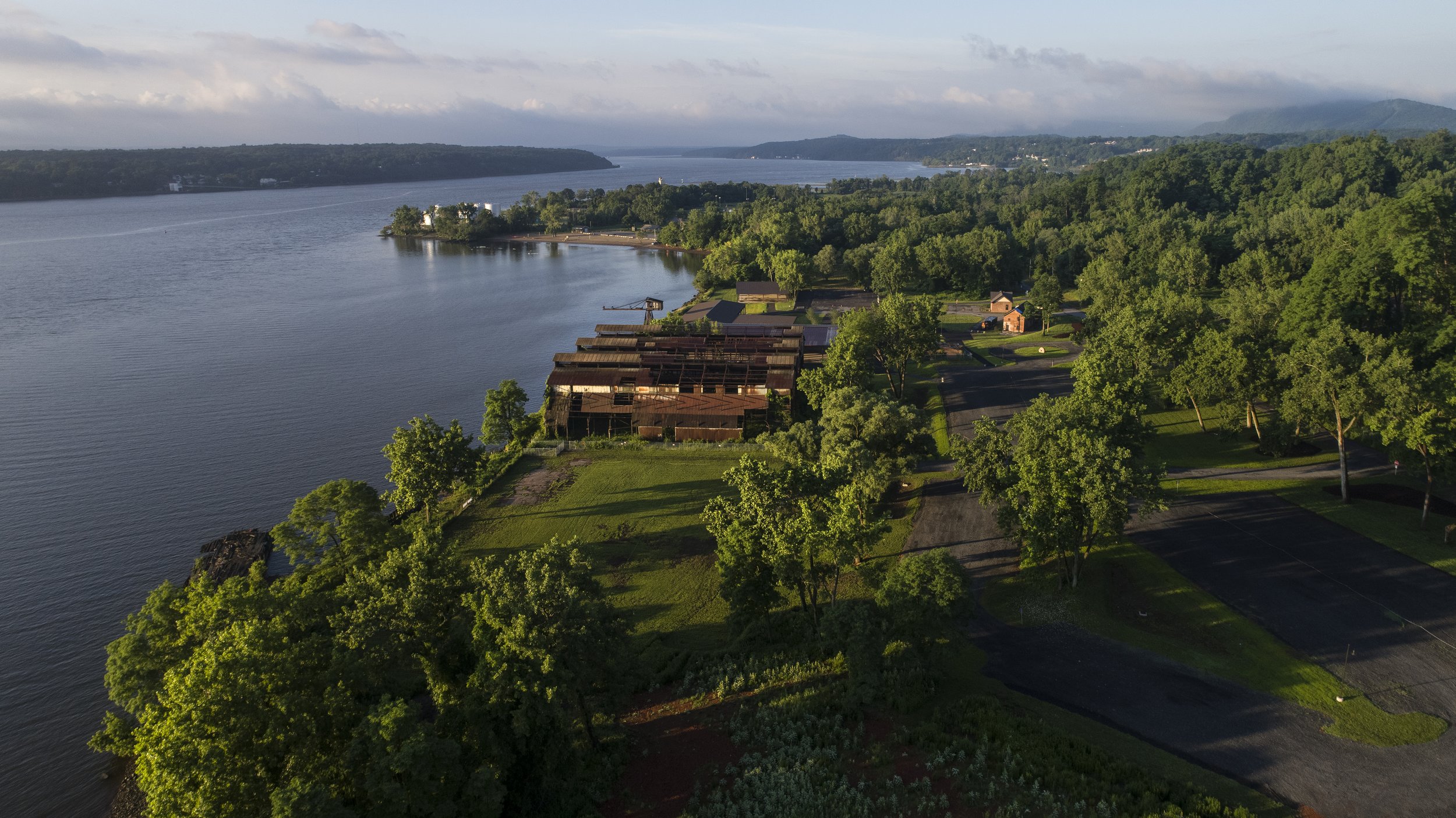 Aerial view of a river with green trees lining the shore, some old wooden structures, and parking lots, with hills and cloudy sky in the background.
