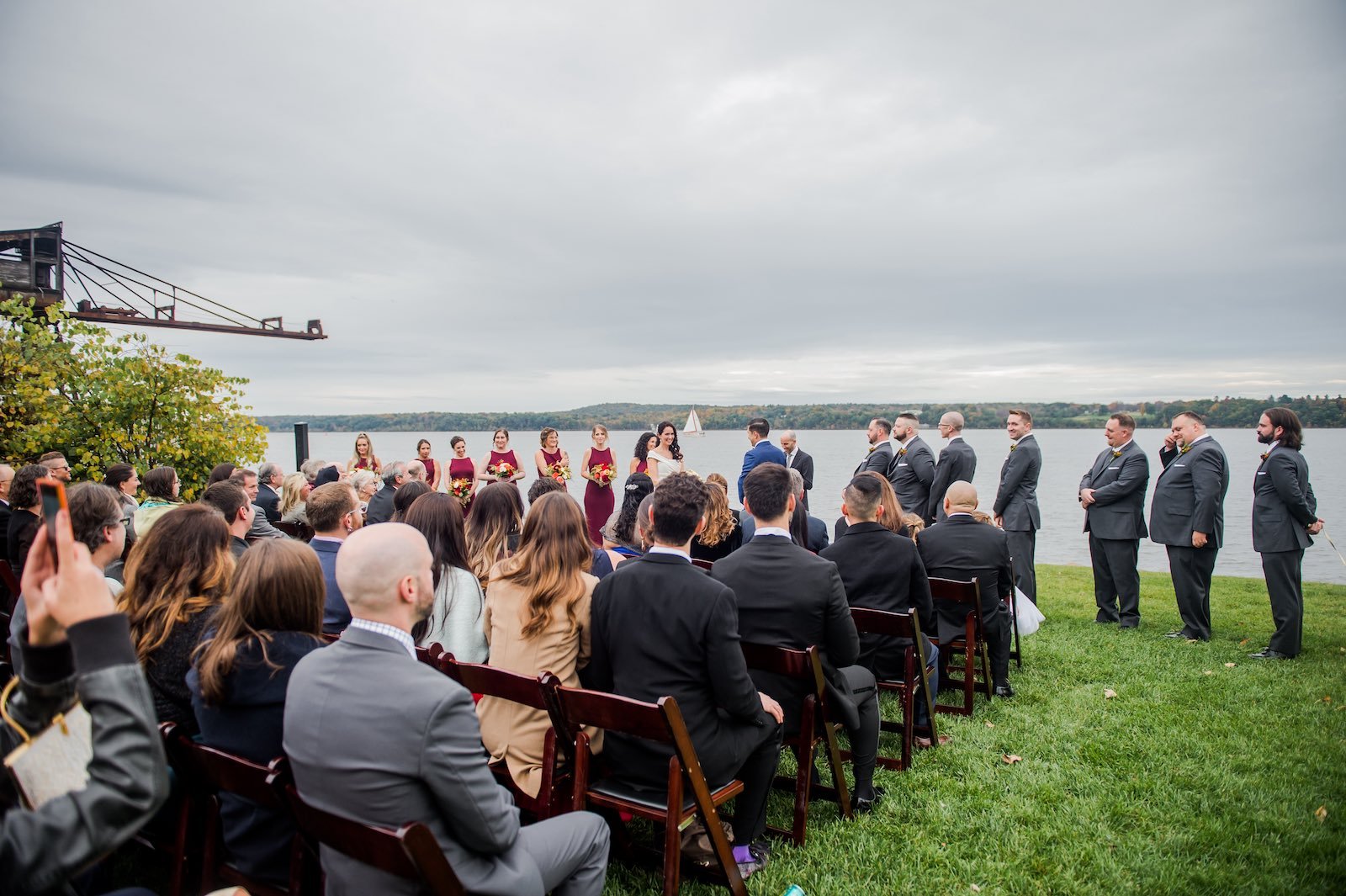 A wedding ceremony taking place outdoors by a body of water on a cloudy day, with the bride and groom standing in front of a seated crowd and a bridal party in matching dresses and suits.