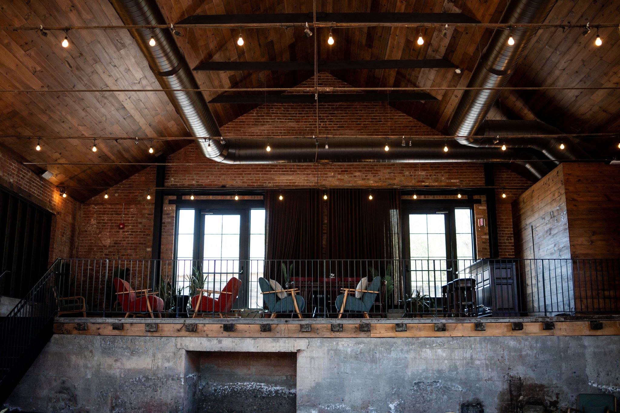 Interior of a loft-style restaurant or lounge with brick walls, wooden ceiling, string lights, and a small elevated balcony with colorful chairs and plants.