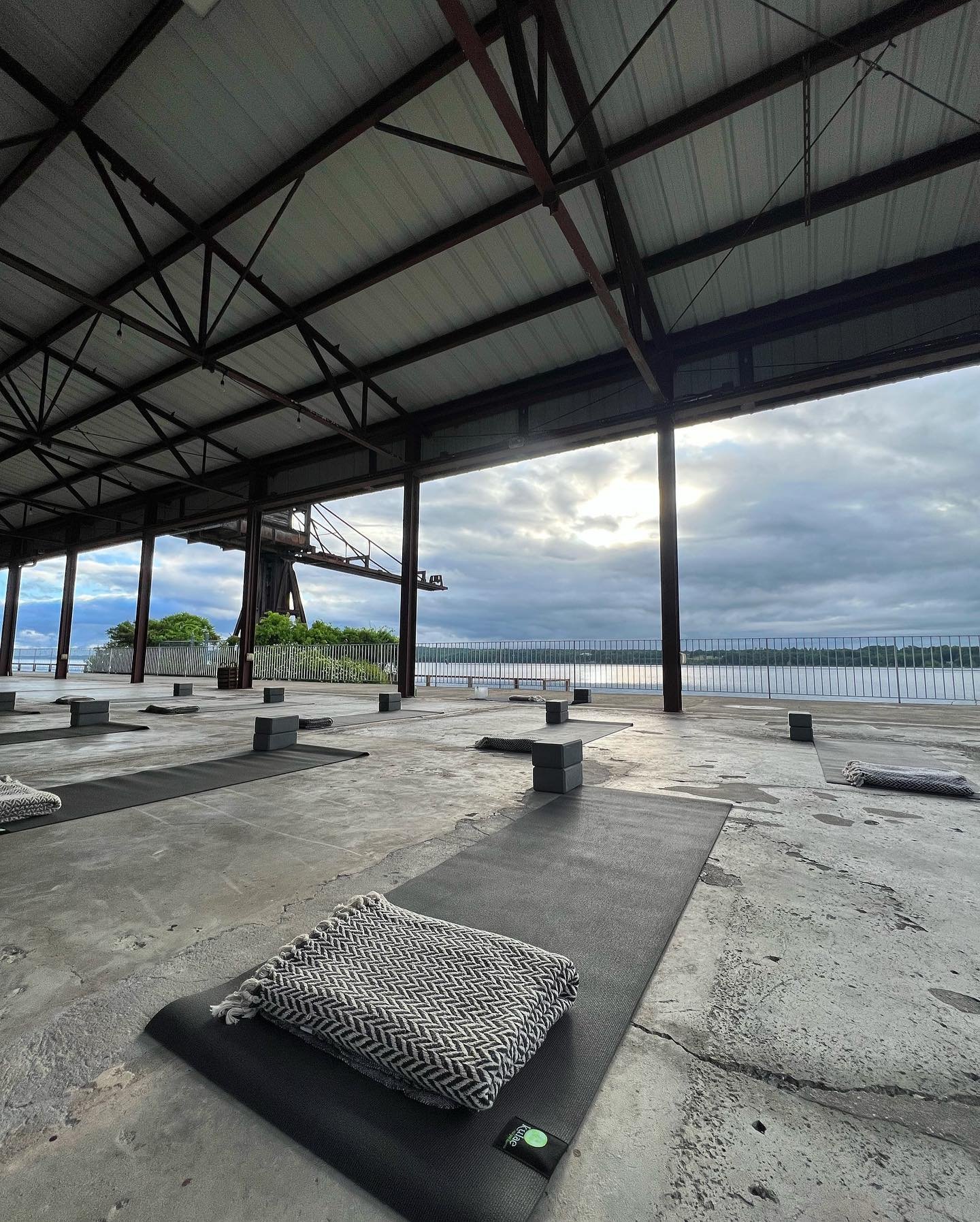 An empty outdoor yoga deck near a river with black mats and stacked blankets, overlooking a cloudy sky and greenery in the background.