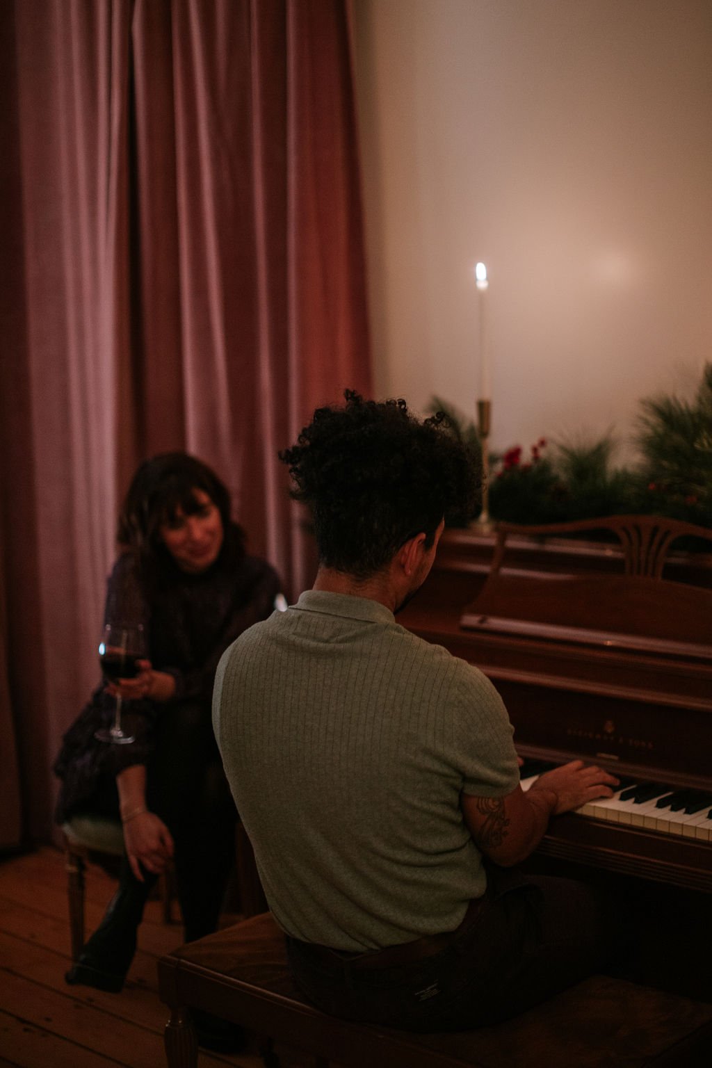 A man with short curly hair and tattooed arms playing a piano indoors, while a woman with dark hair holding a glass of wine watches.