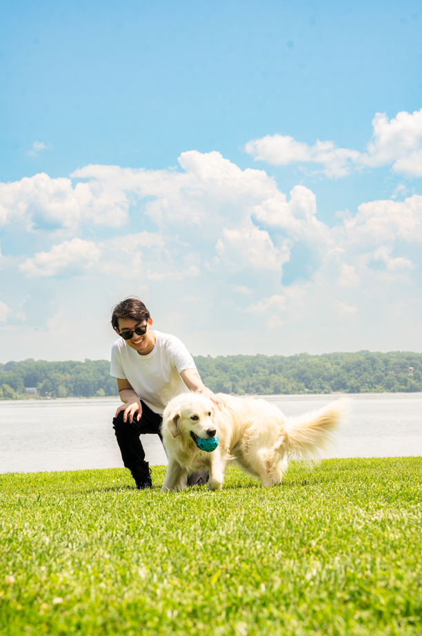 A woman with sunglasses playing with a golden retriever dog on a grassy area near a lake, with a blue sky and fluffy clouds in the background.