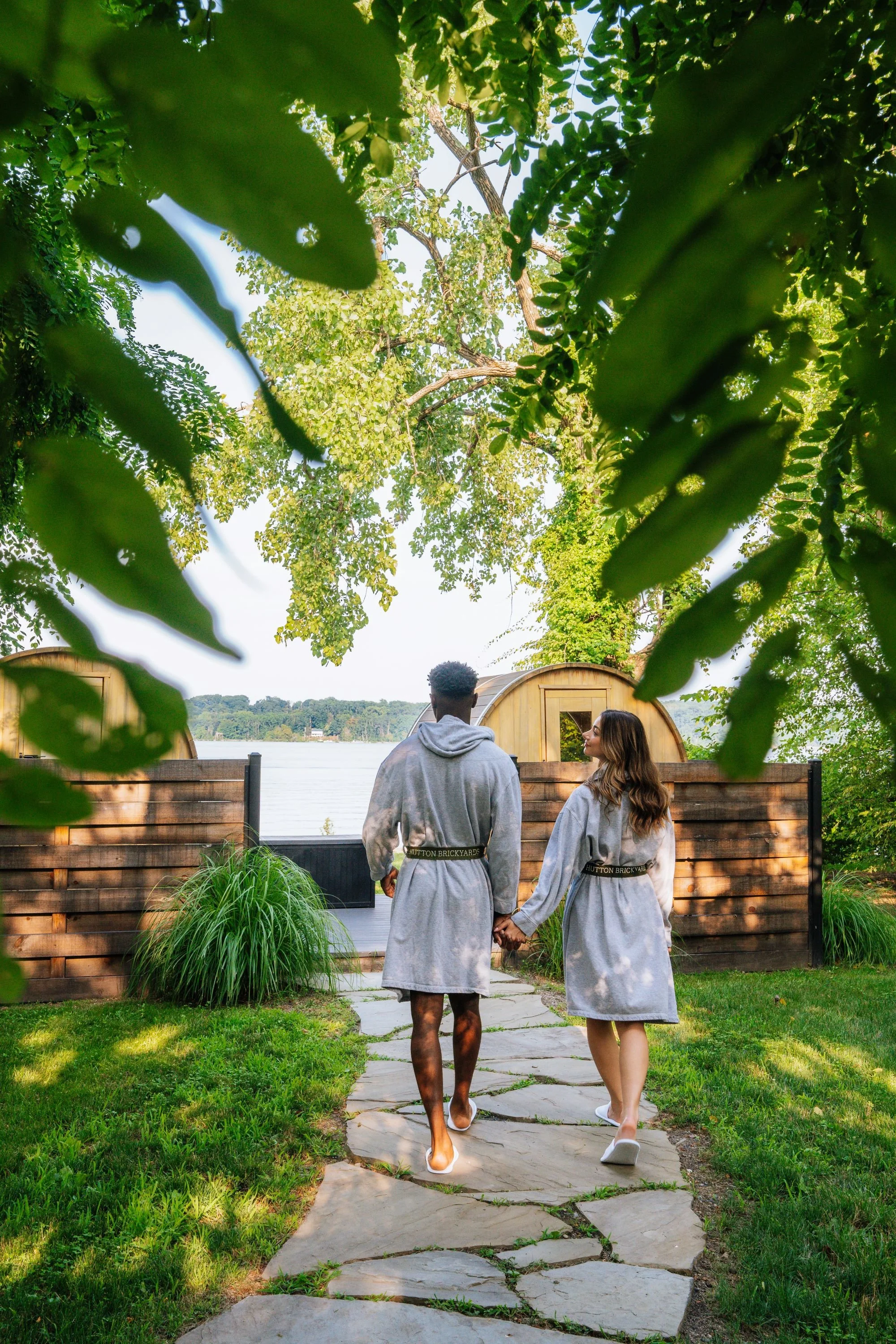 A couple dressed in grey robes holding hands, walking on the spa garden stone path towards the waterfront saunas, surrounded by green grass and trees.