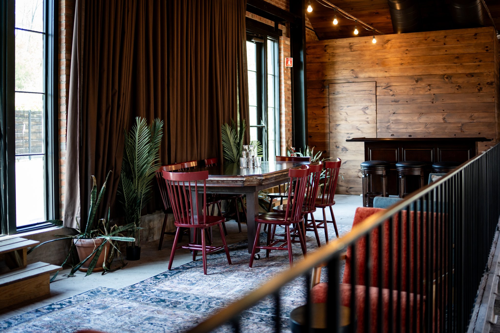 Interior of a cozy restaurant or cafe with large windows, brown curtains, a wooden wall, a round table surrounded by red chairs, potted plants, a bar with black stools, and warm lighting.