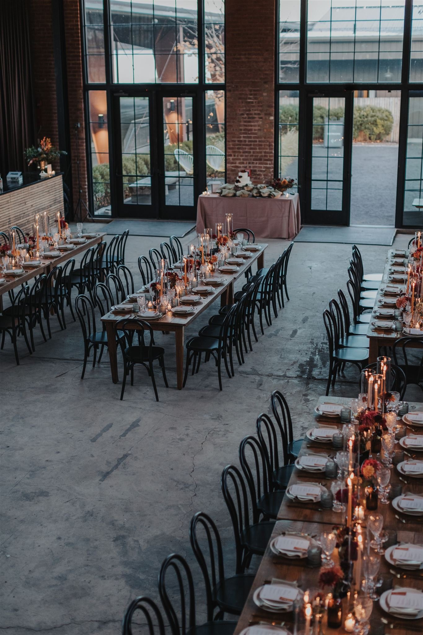 Interior of a decorated event space with long dining tables set with candles, flowers, dinnerware, and chairs, large windows, and natural light.