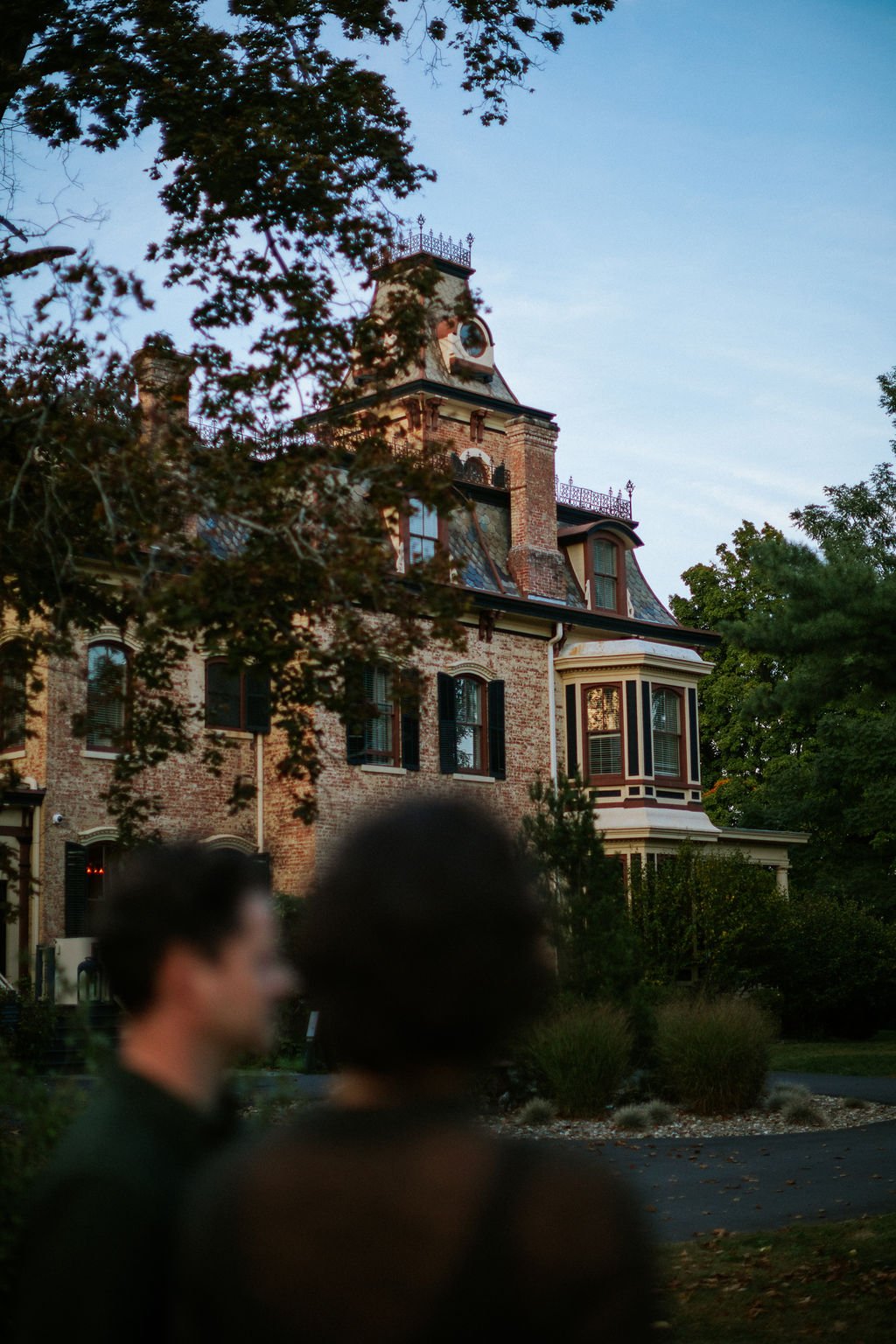 A large, historic brick mansion with Victorian architecture, surrounded by trees, with two blurred people walking in the foreground.