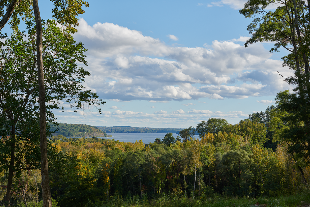 Scenic view of the Hudson River, surrounded by green trees and a partly cloudy sky.