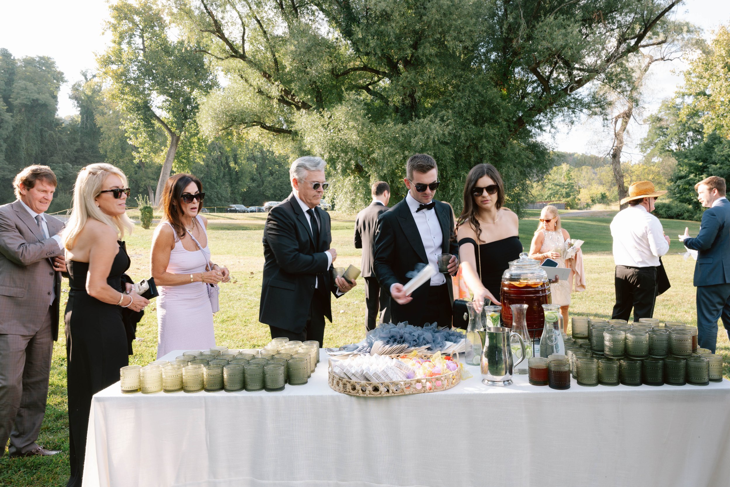 People in formal attire at an outdoor event gathering around a table with drinks and snacks, set in a park with large trees and green grass.