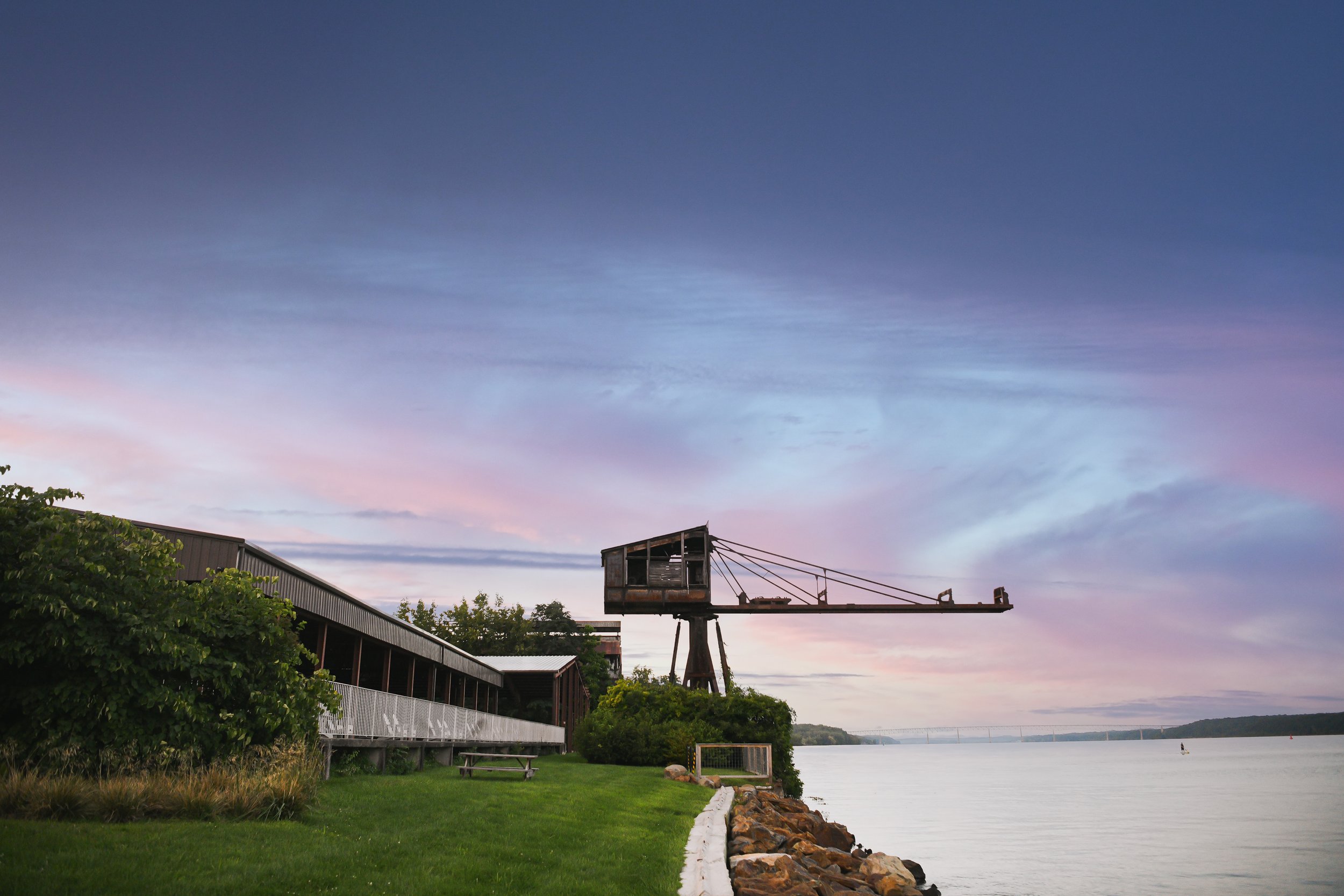 A rustic old crane over a body of water with a grassy shoreline and a building to the left at sunset or sunrise with a colorful sky.