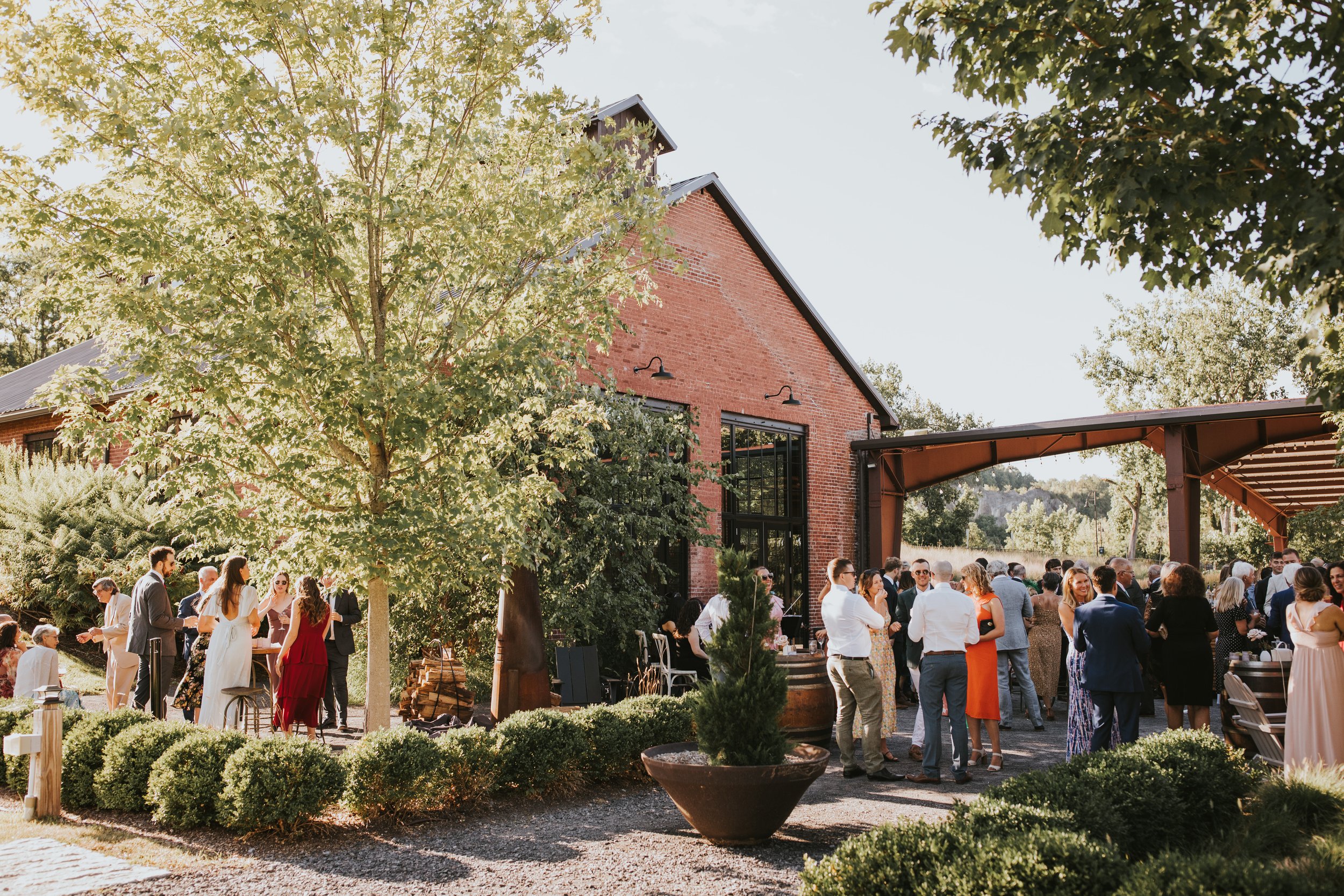 Outdoor gathering of people socializing near a brick building with large windows, surrounded by greenery and trees, during daytime.