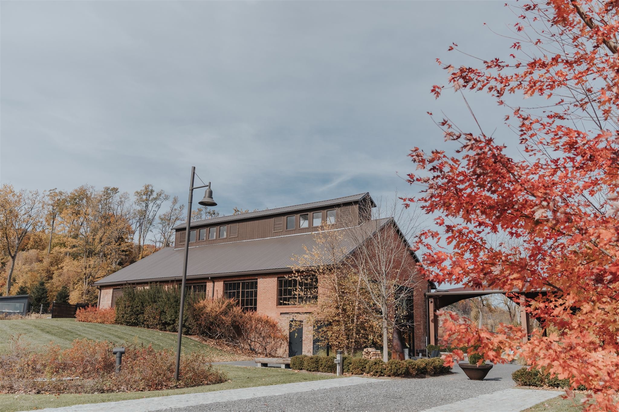 A large brick building with a metal roof and multiple windows, surrounded by autumn trees with orange and yellow leaves, a neatly trimmed lawn, a gravel pathway, and a lamppost under a cloudy sky.