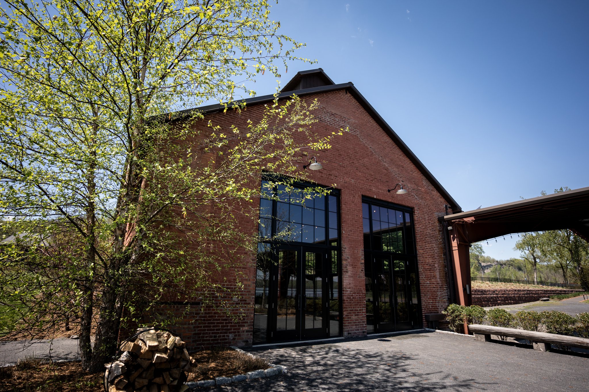 A brick building with large black-framed windows under a clear blue sky, surrounded by trees and outdoor seating.