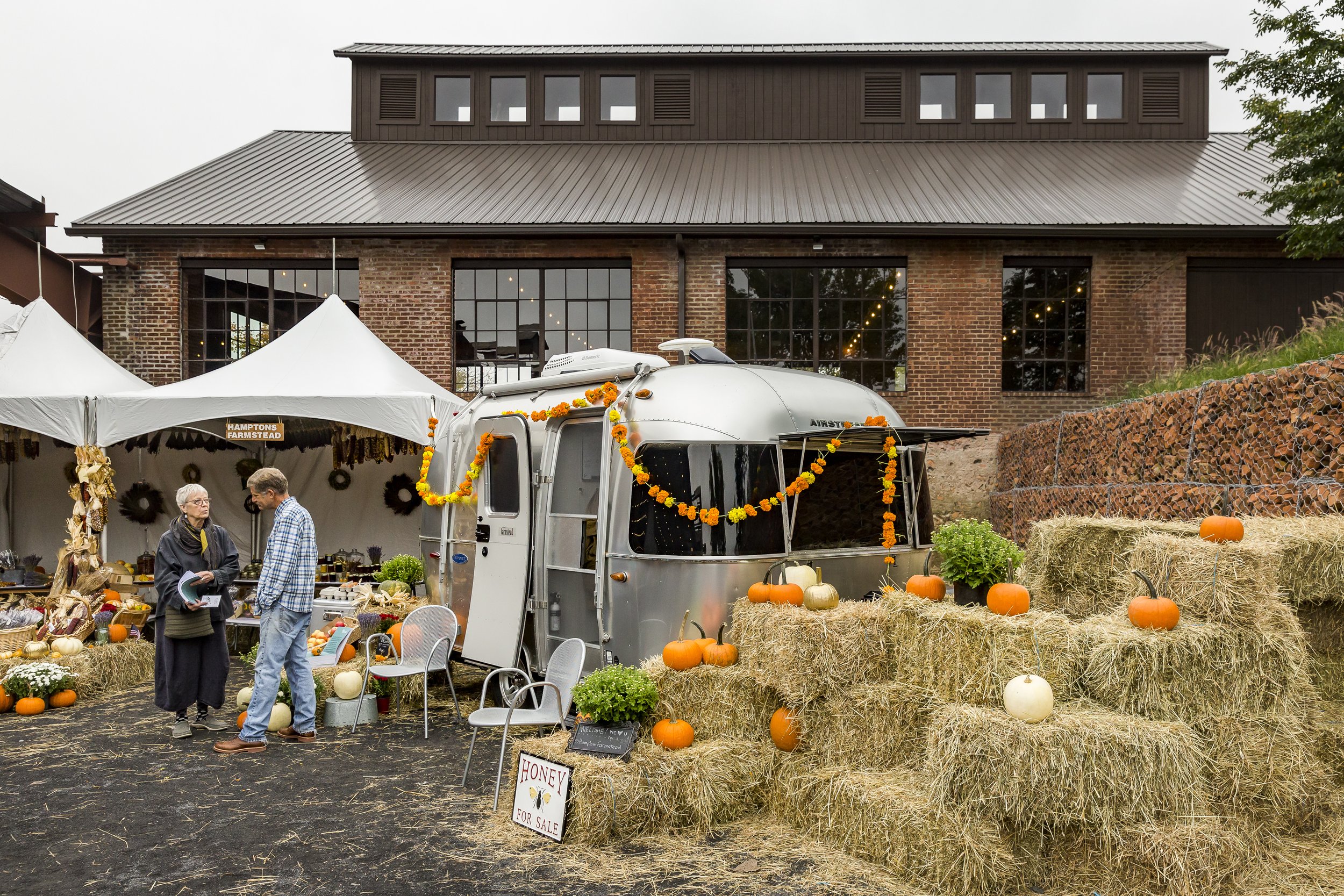 An outdoor farmer's market stall decorated with pumpkin and flower garlands, hay bales, and a vintage silver trailer with Halloween decorations in front. Two people are conversing in front of the stall, and the background shows a brick building with