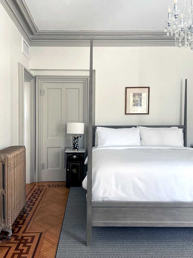 A bedroom with a gray wooden four-poster bed, white bedding, a nightstand with a lamp, a framed picture on the wall, a vintage radiator, a door, and a chandelier.