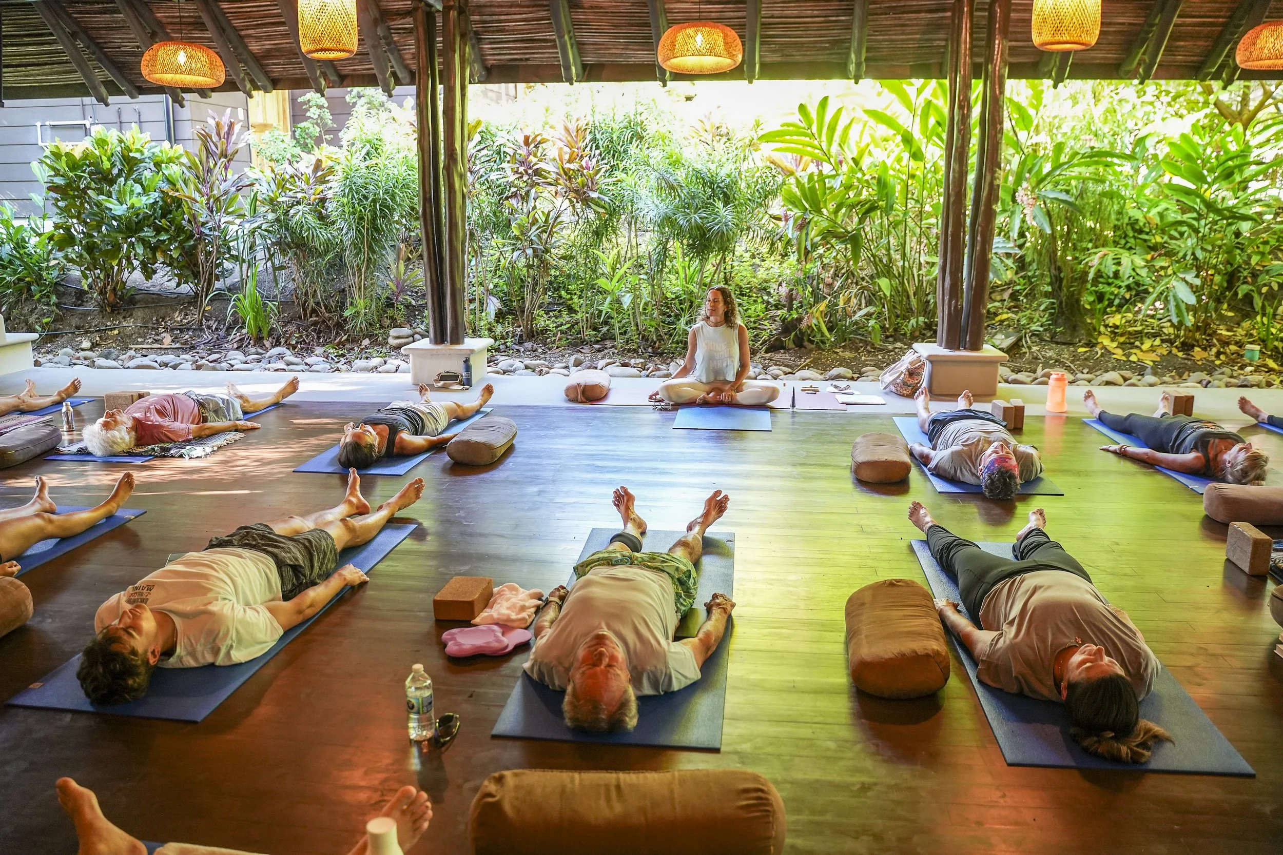 Yoga teacher guiding a class in savasana in an open-air shala surrounded by tropical gardens