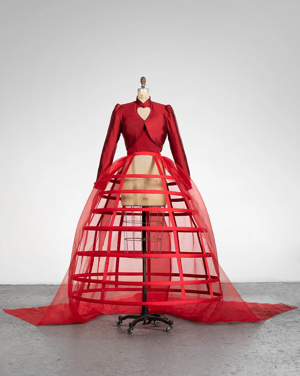 A mannequin dressed in a red fashion outfit with a fabric cage skirt and a jacket with a heart-shaped cutout on the chest, displayed in a plain gallery room.