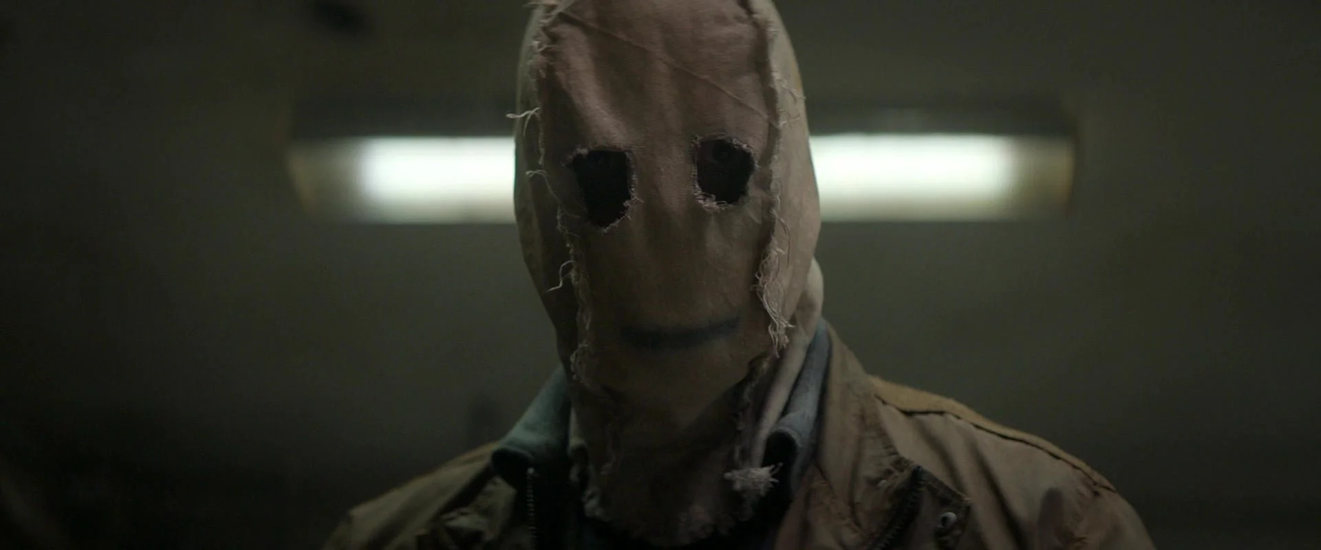 Person wearing a tattered, distressed mask with holes for eyes, standing in a dimly lit room.