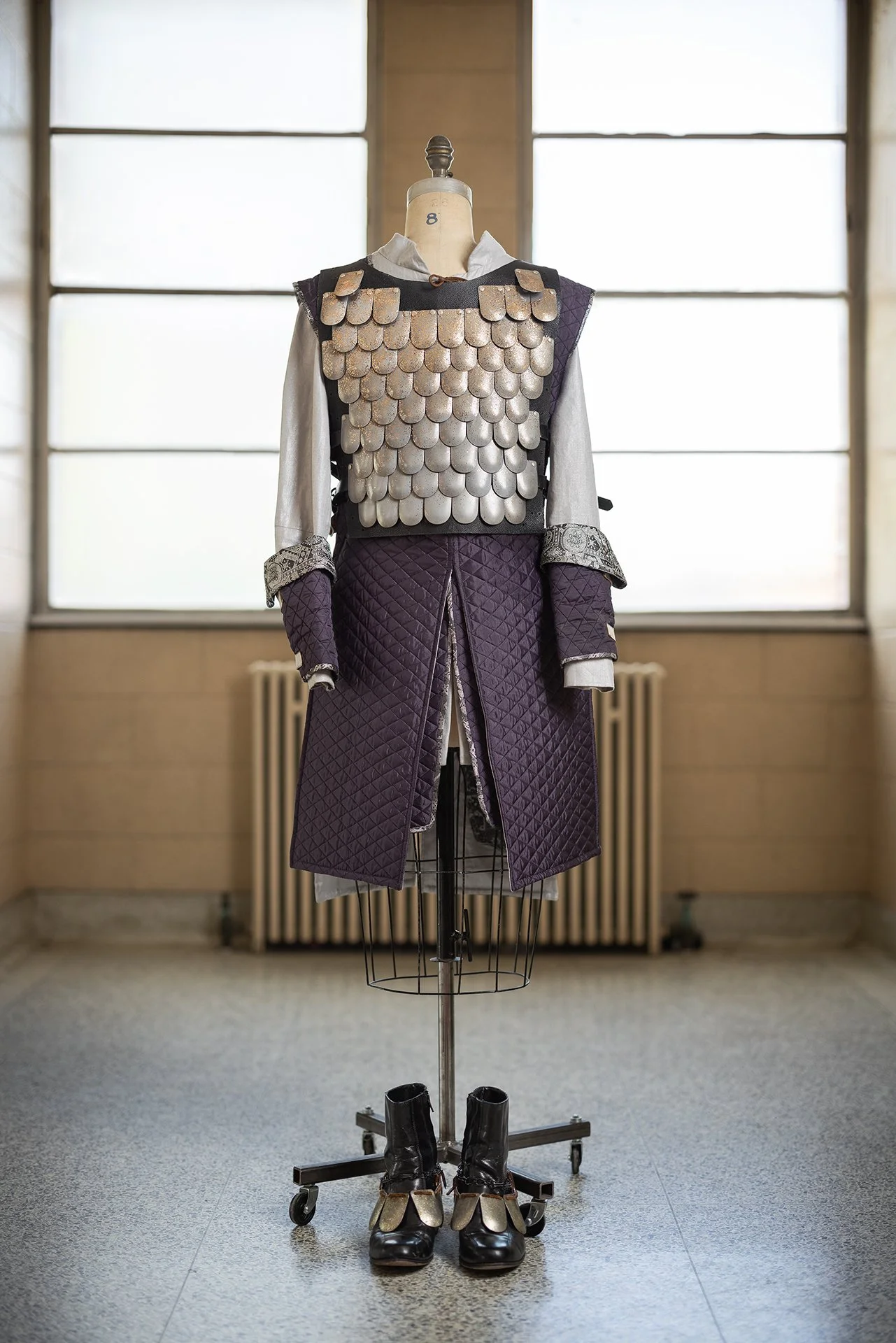 Vintage costume display featuring a metal armor chest piece, quilted purple garment, and matching sleeves, with boots and shoes on the floor, set in front of large windows.