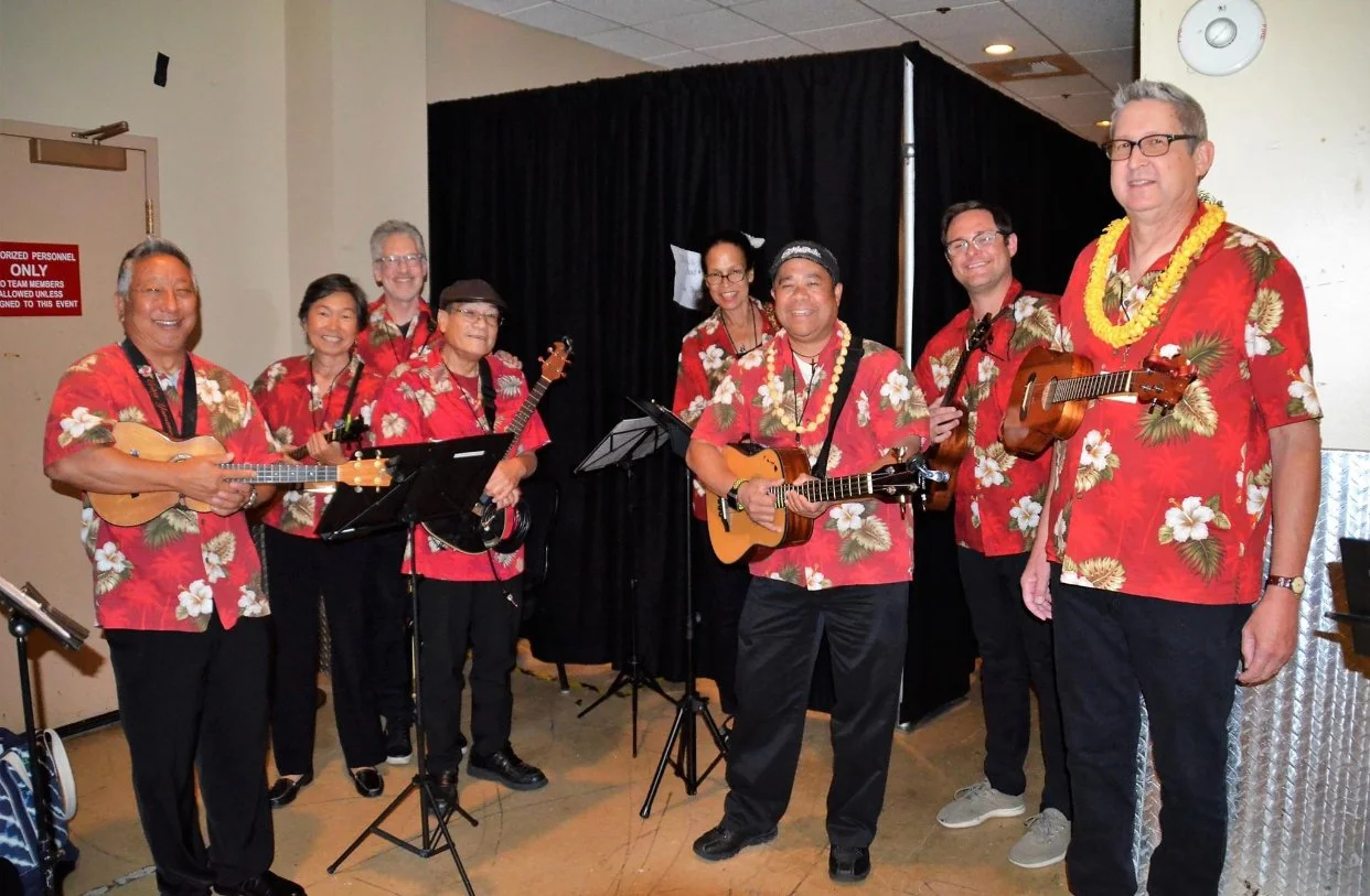 Group of musicians in Hawaiian shirts holding ukuleles and guitars, standing indoors.