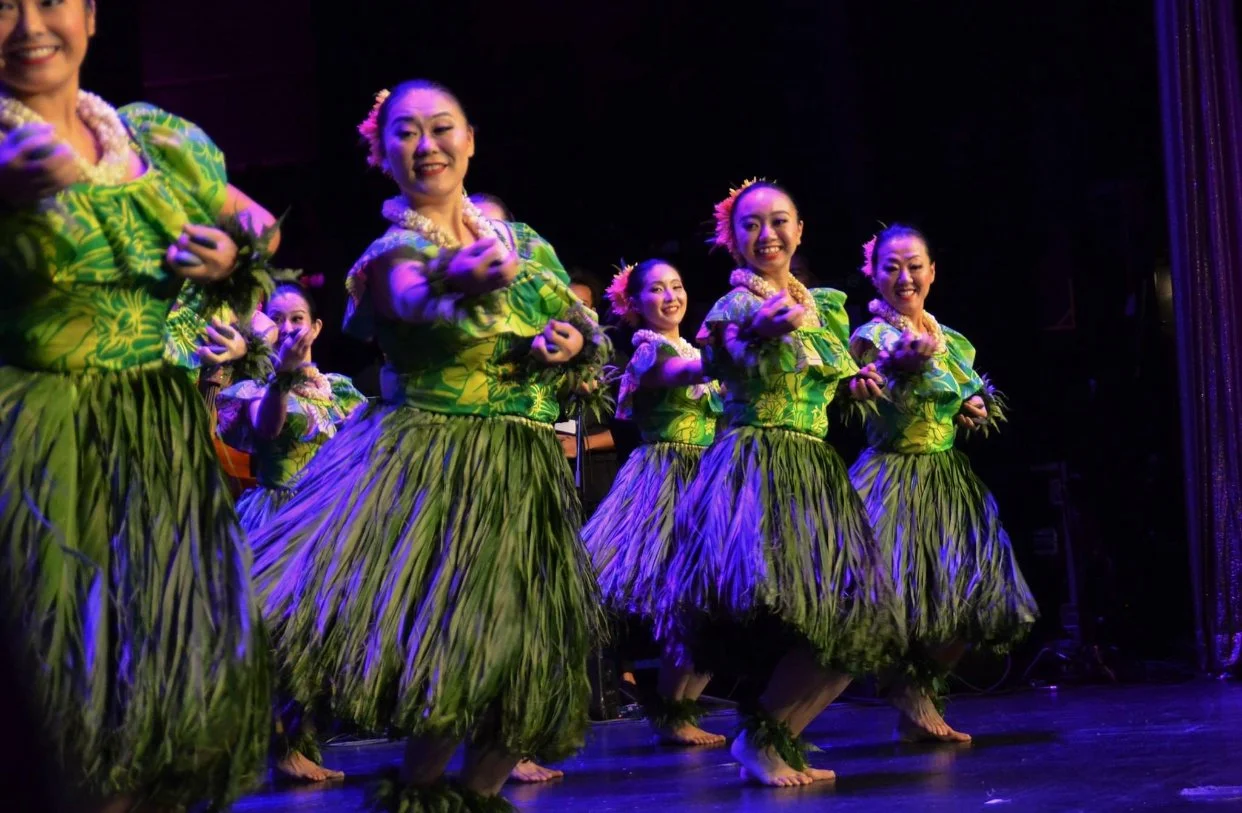 Group of dancers performing in traditional Hawaiian hula attire with green skirts and floral necklaces on stage.