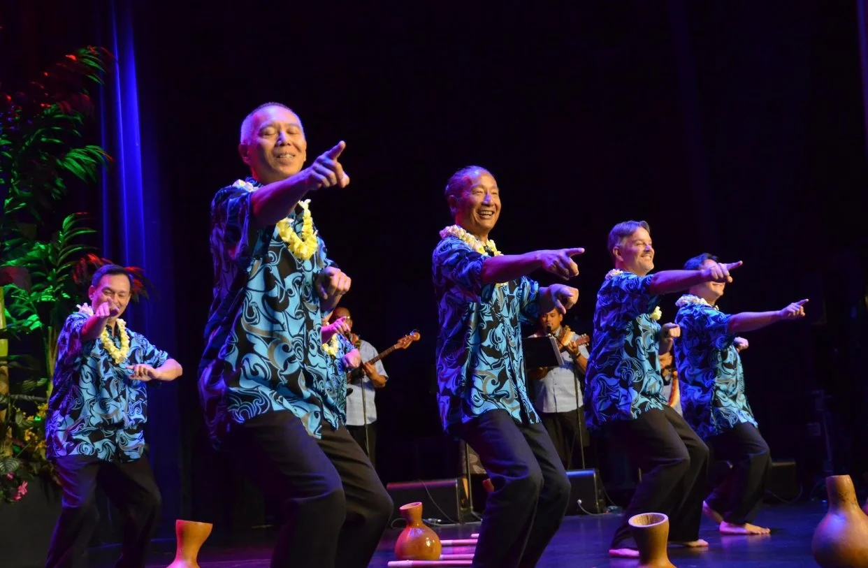Group of men in blue Hawaiian shirts performing a dance on stage with musicians playing in the background.
