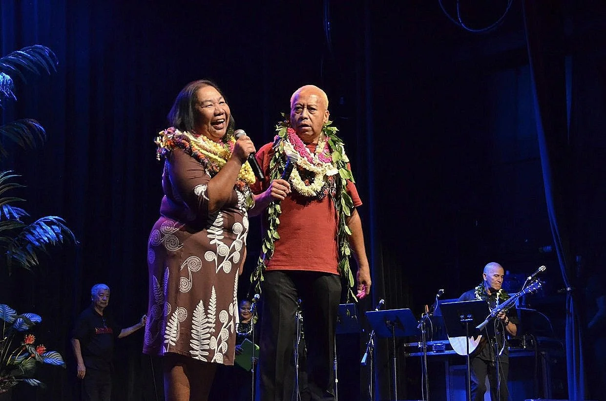 Two people wearing leis sing on stage, one holding a microphone, with musicians in the background.